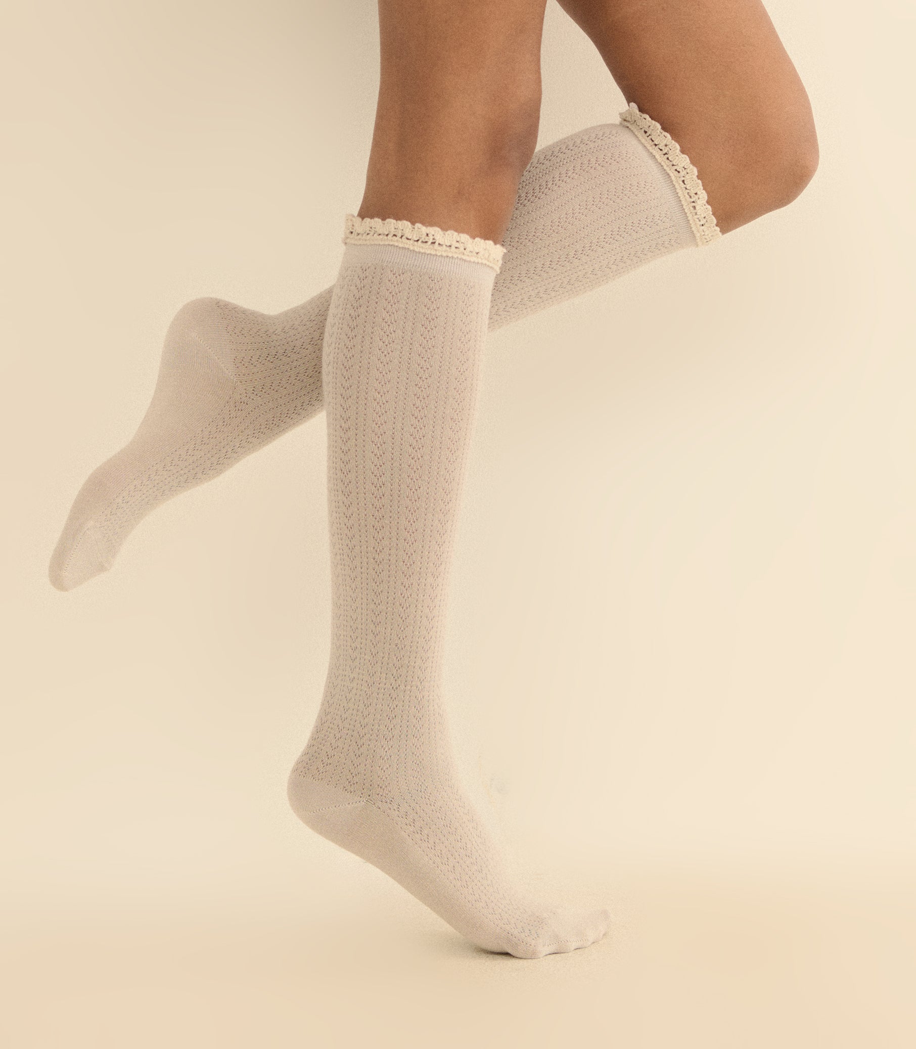 A pair of cream-colored knee-high socks with a cable knit pattern and a delicate lace trim at the top, shown on a model's legs against a warm beige background.