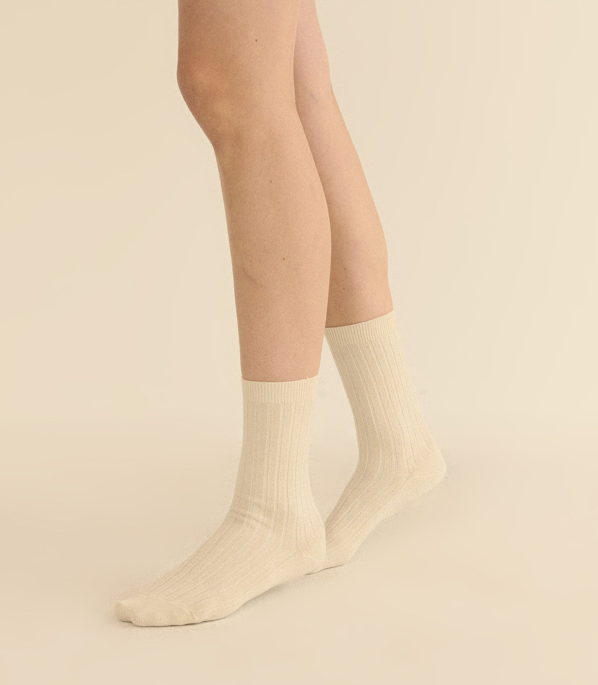 A pair of ribbed crew socks in cream beige, worn on a model's feet against a solid light beige background. 1
