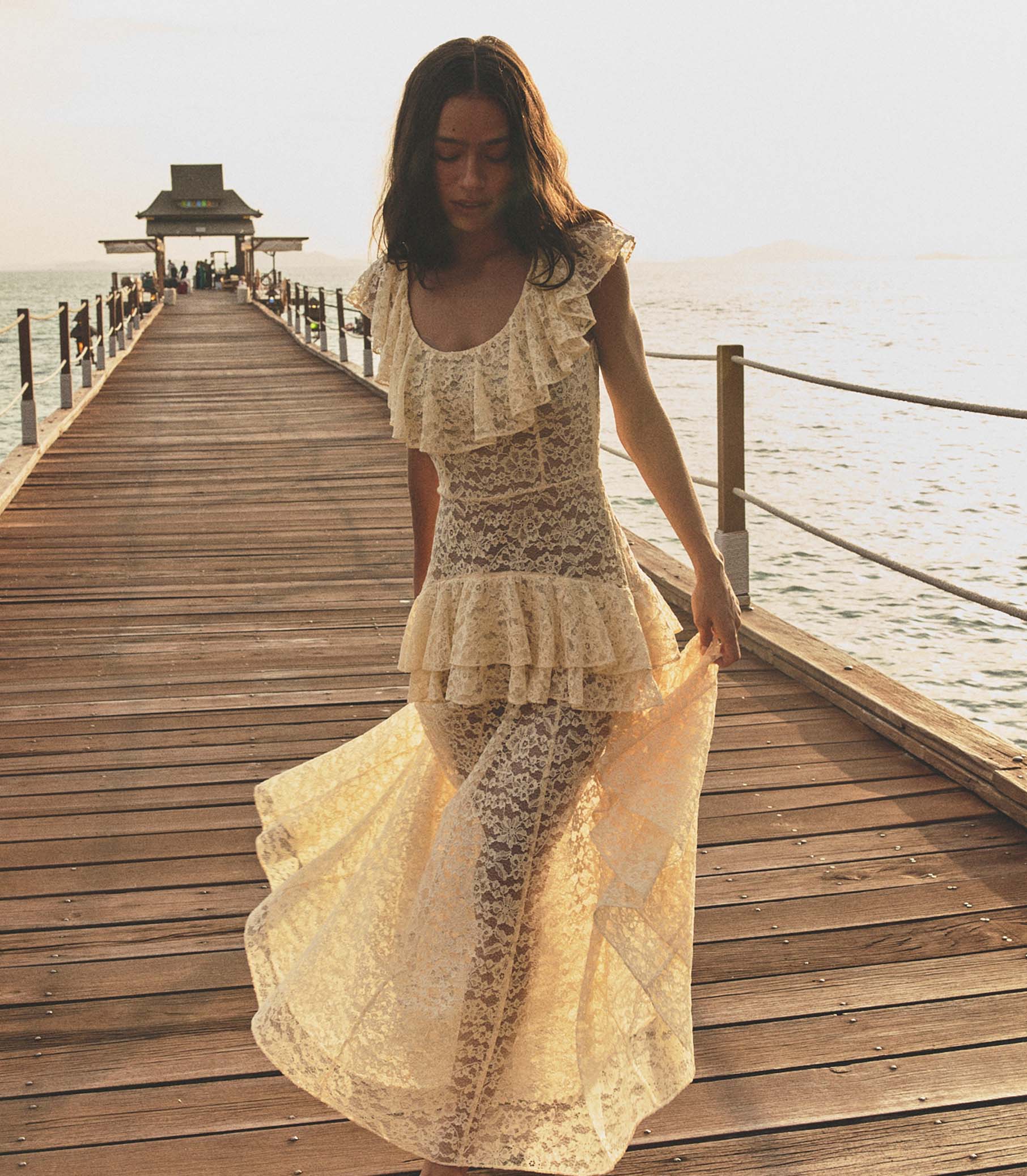 Woman in a cream lace dress walking on a wooden pier by the water.