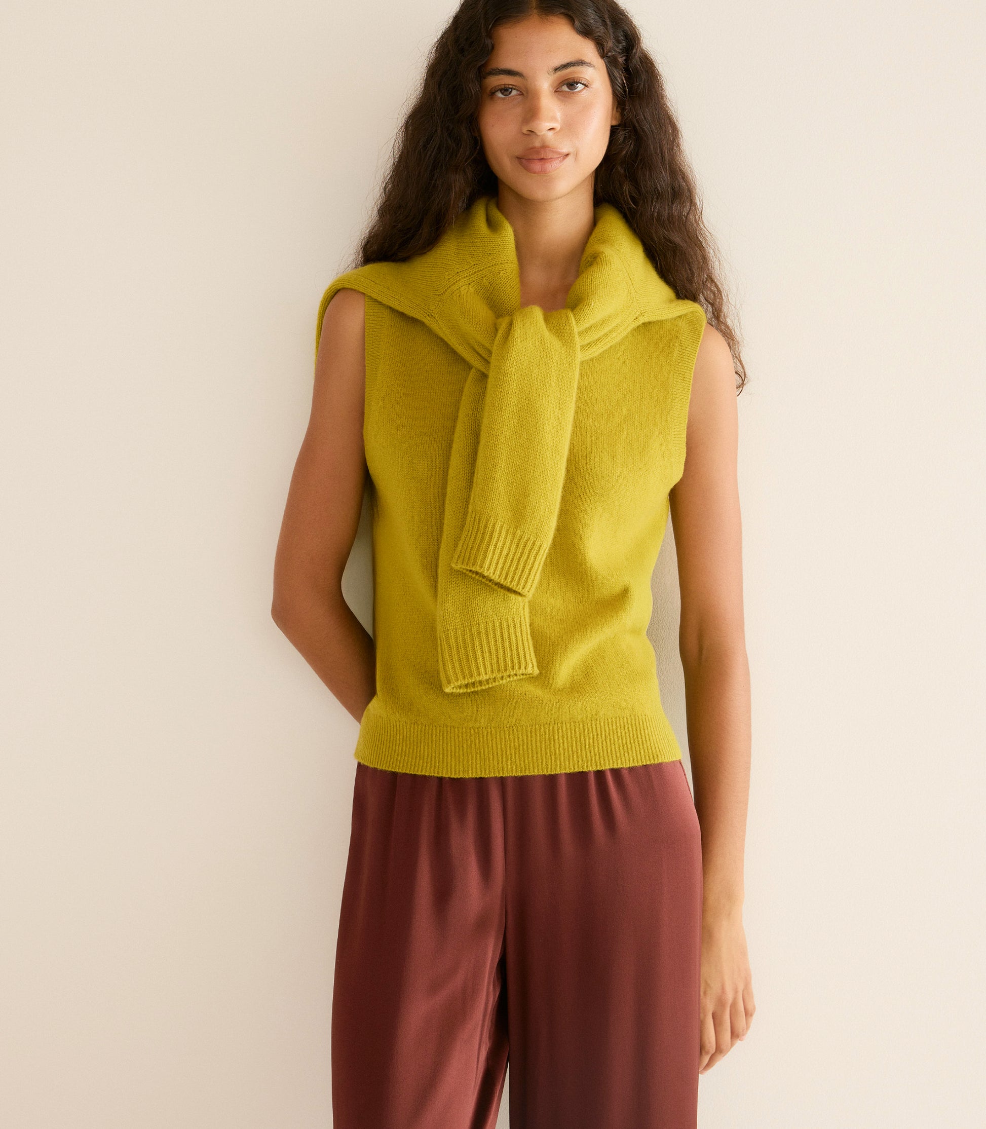 Woman wearing a mustard yellow sleeveless sweater with a matching scarf against a beige background 3
