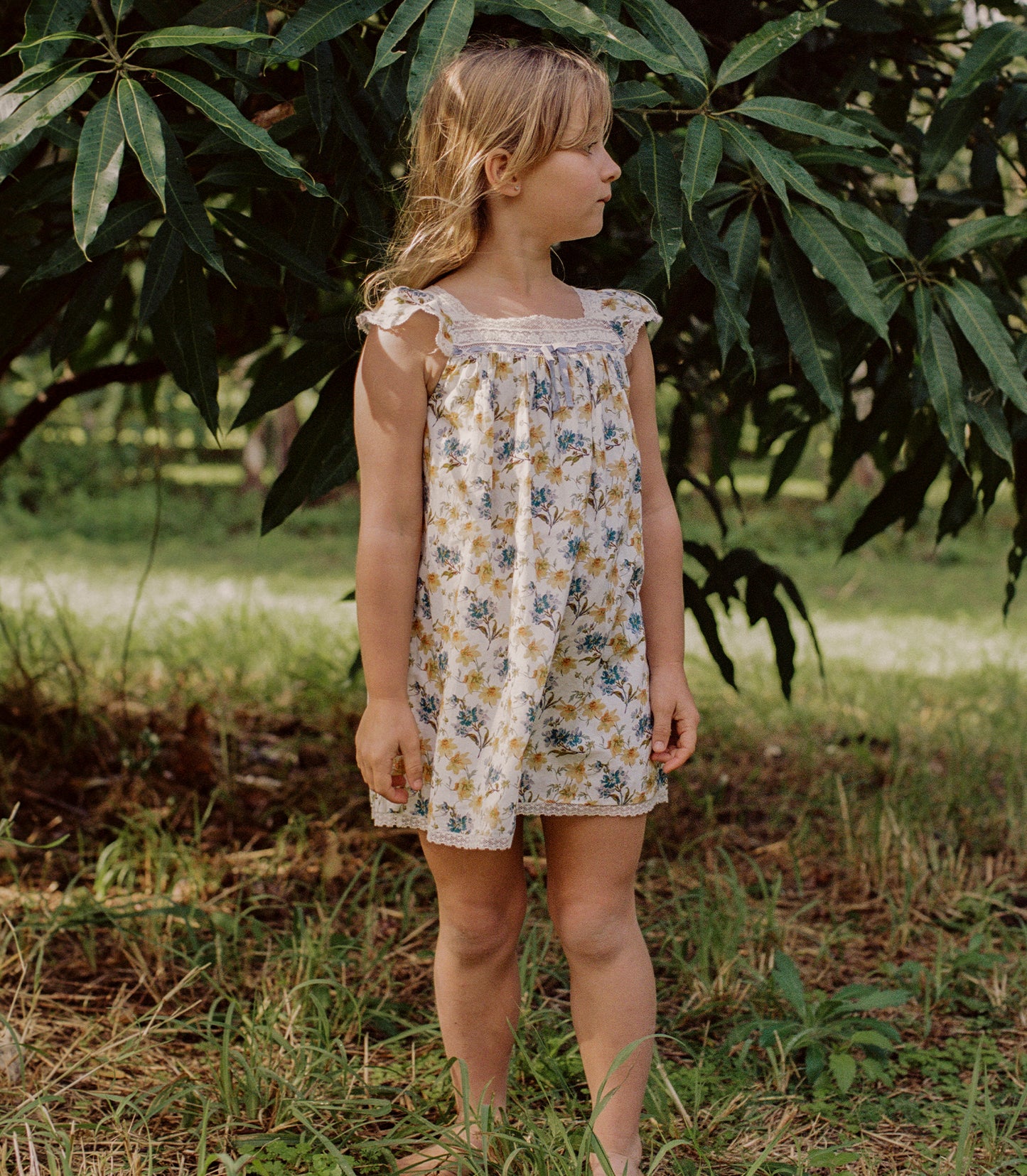 A young girl wearing a sleeveless cream-colored dress with a blue and yellow floral print, standing outdoors in a grassy area under leafy green trees.