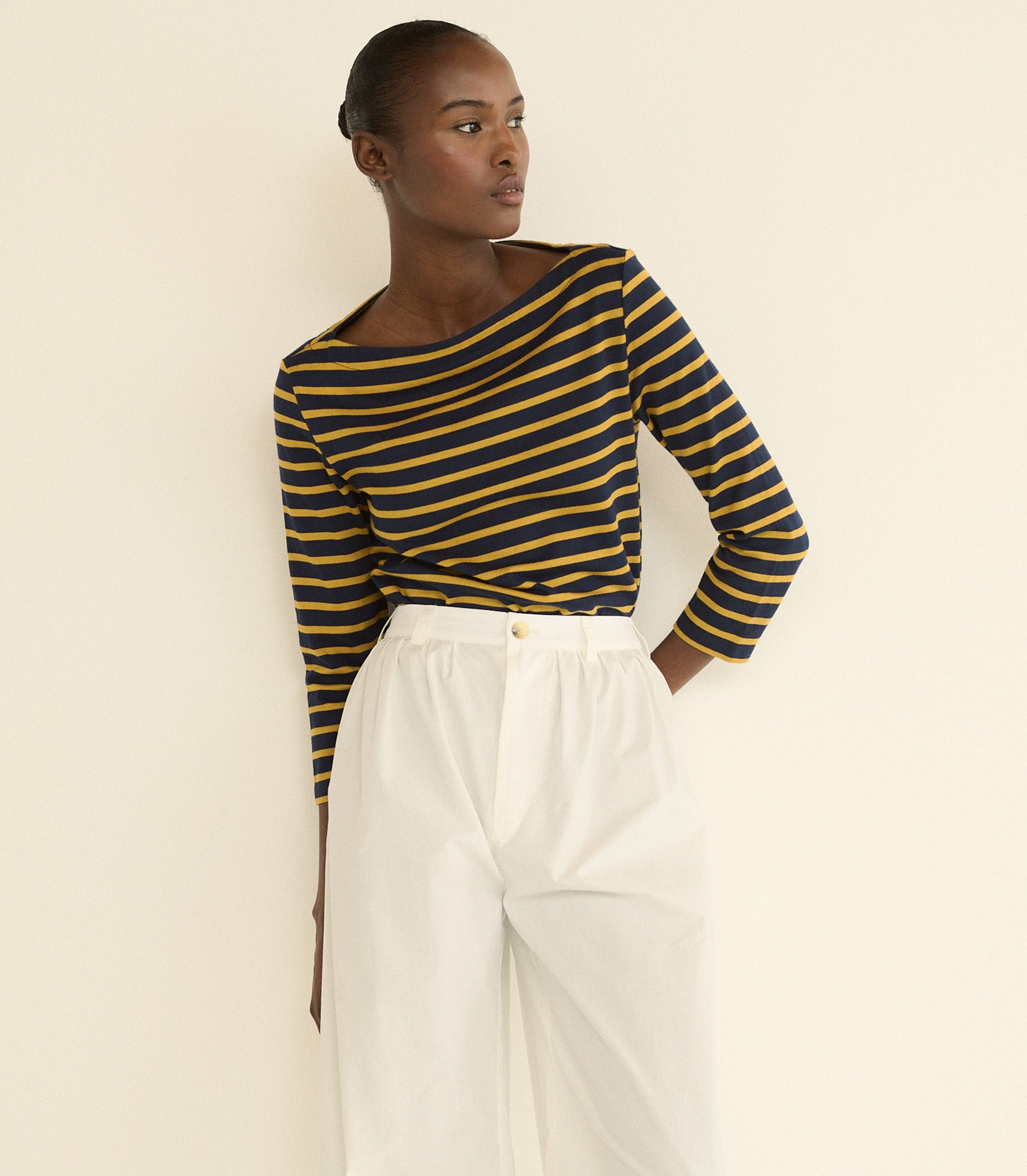 A model wearing a navy and mustard yellow striped long-sleeve boat-neck top paired with white high-waisted trousers, standing against a plain cream-colored wall. 1
