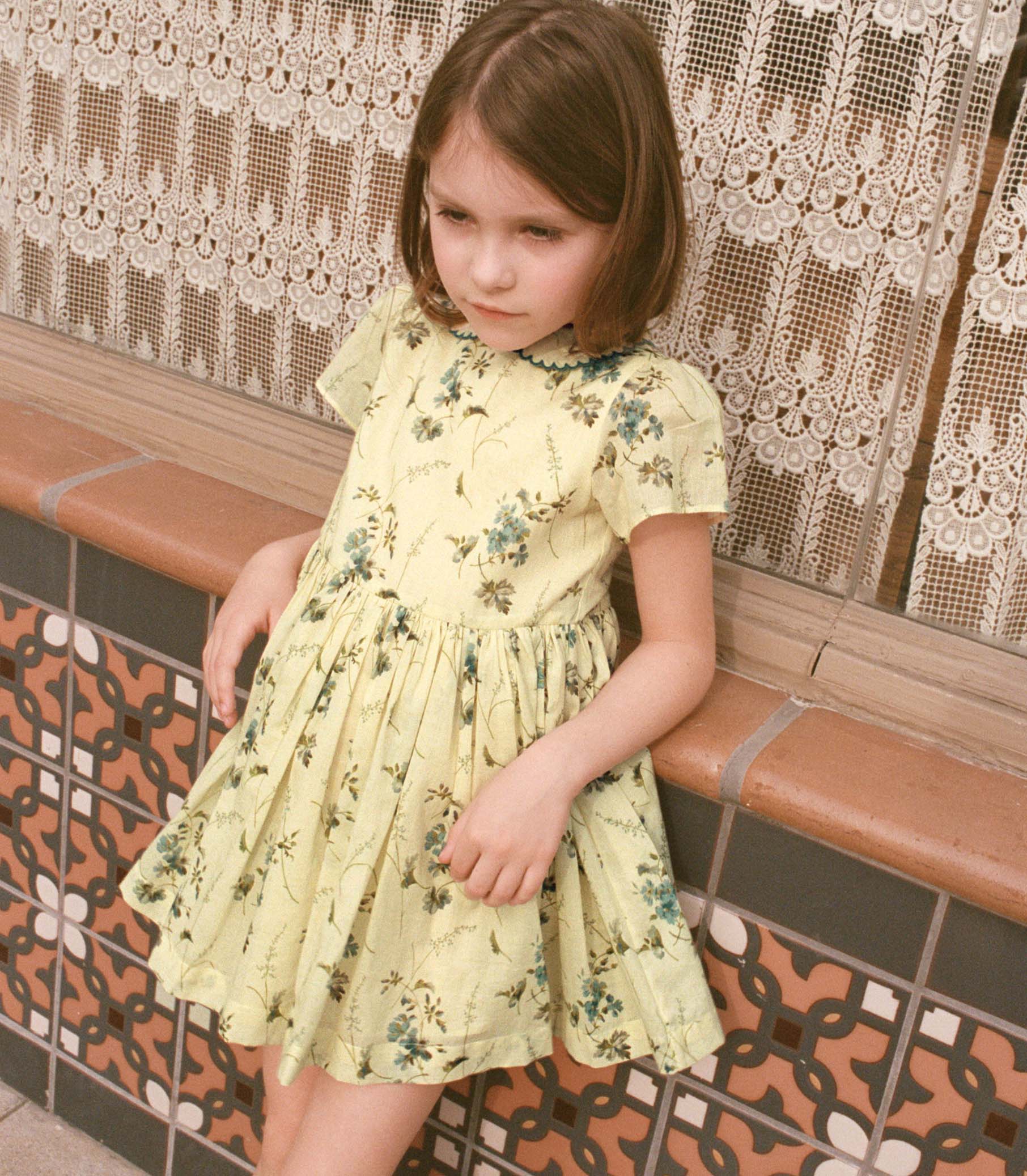 Young girl in a yellow floral dress sitting on a tiled bench with lace curtains in the background. 1

