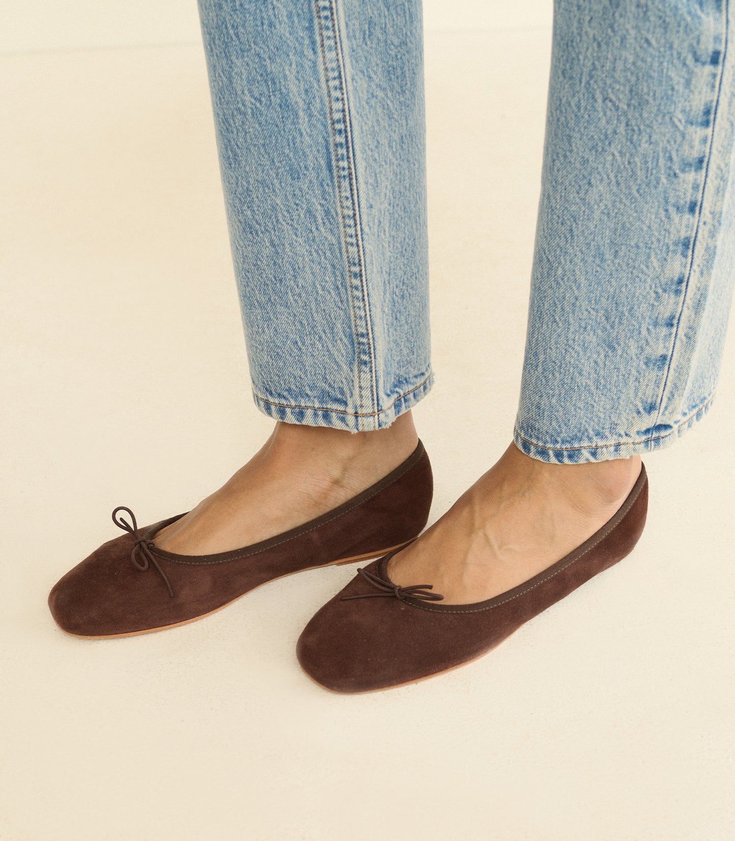 A person wearing dark brown suede ballet flats with small bow details, paired with light-wash blue jeans, shown from the calves down against a plain light beige background.