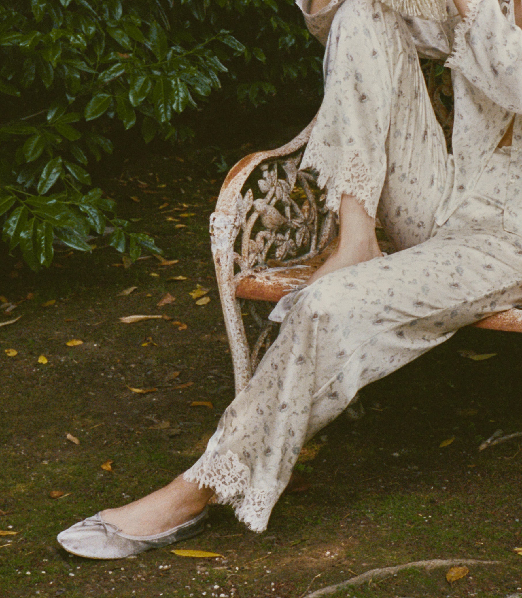 A person seated on an outdoor garden bench wearing a loose cream-colored silk pants with lace trim and light flats, surrounded by green foliage and a dirt ground. 1
