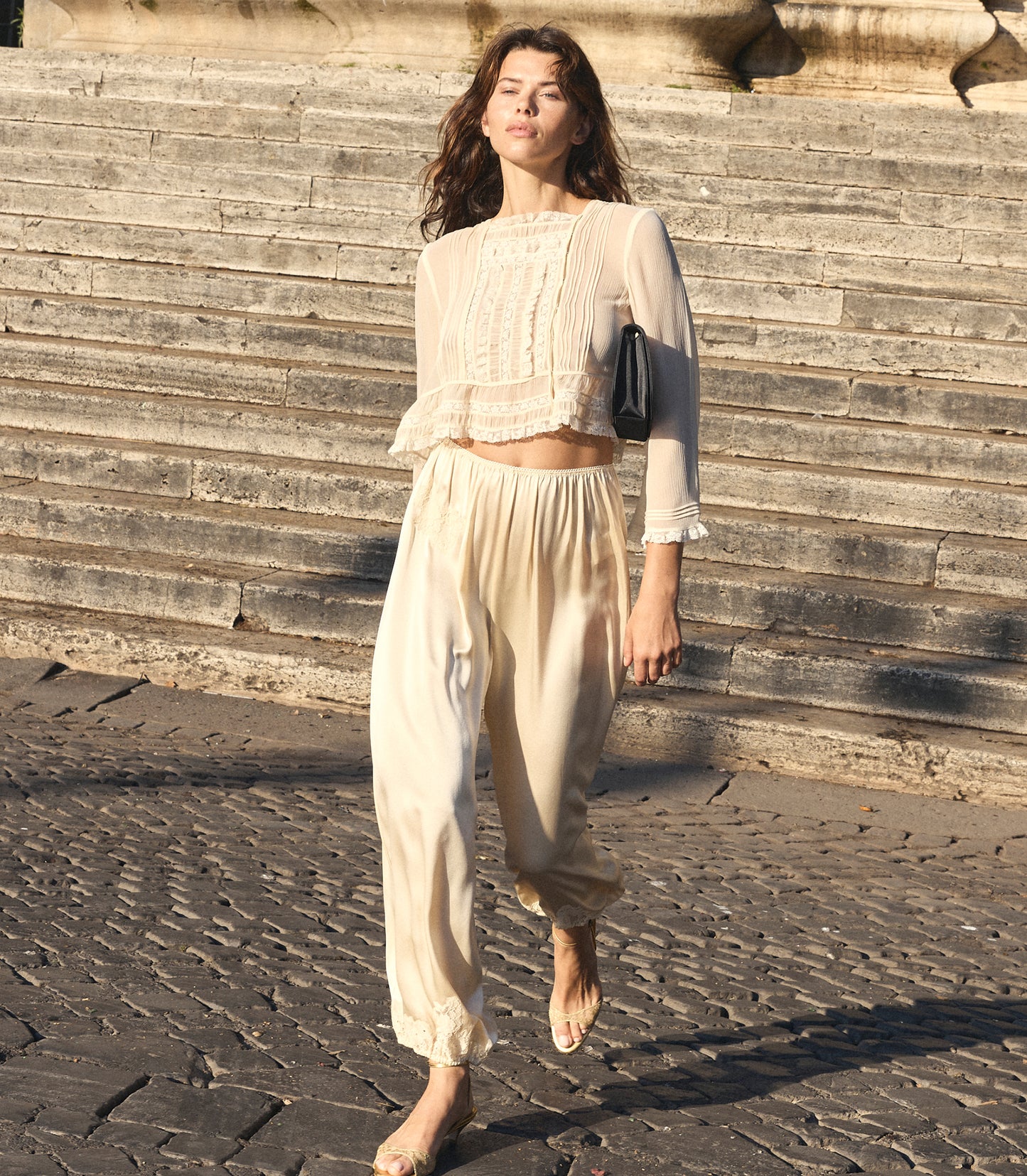 A woman walking outdoors on stone steps wearing a cream-colored two-piece outfit consisting of a sheer long-sleeve crop top with lace detailing and matching loose trousers. She is carrying a small black shoulder bag.