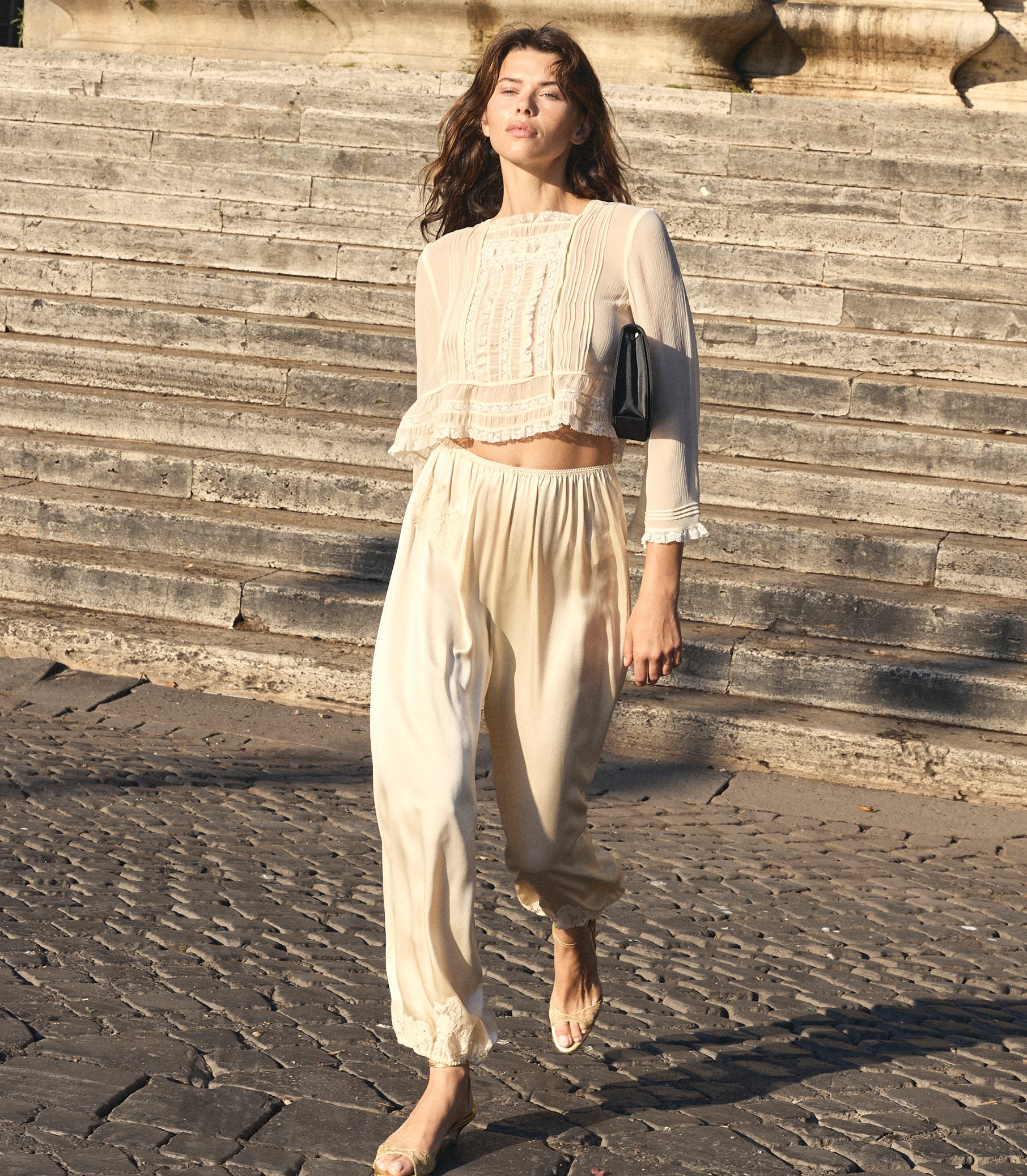 A woman walking outdoors on stone steps wearing a cream-colored two-piece outfit consisting of a sheer long-sleeve crop top with lace detailing and matching loose trousers. She is carrying a small black shoulder bag.