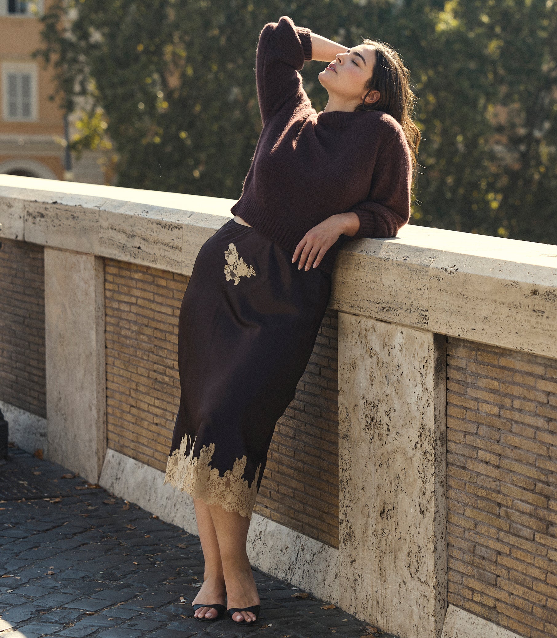 A woman leans against a stone bridge railing outdoors, wearing a dark brown knit sweater and matching midi skirt with beige floral embroidery and a beige lace hem.