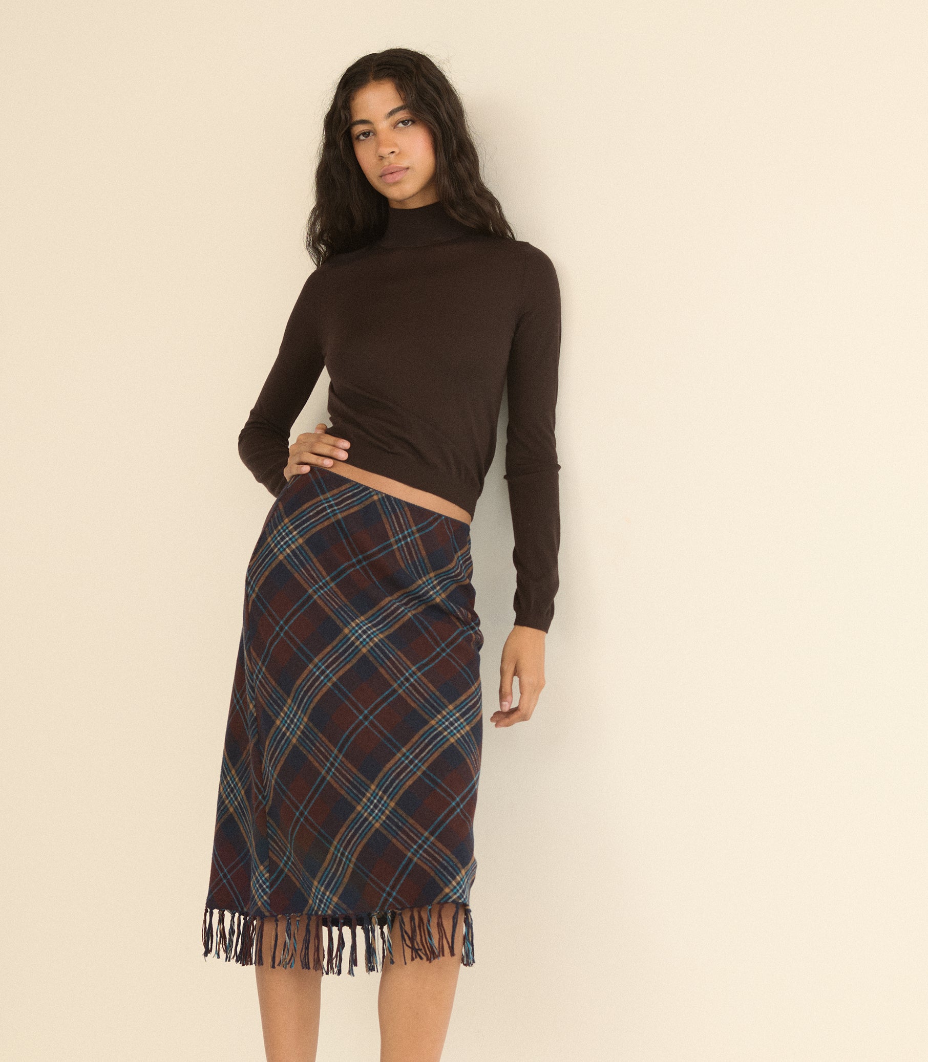 A woman modeling a dark brown long-sleeve mock neck crop top and a dark red and blue tartan plaid wrap skirt with fringe, standing against a plain beige background. 1
