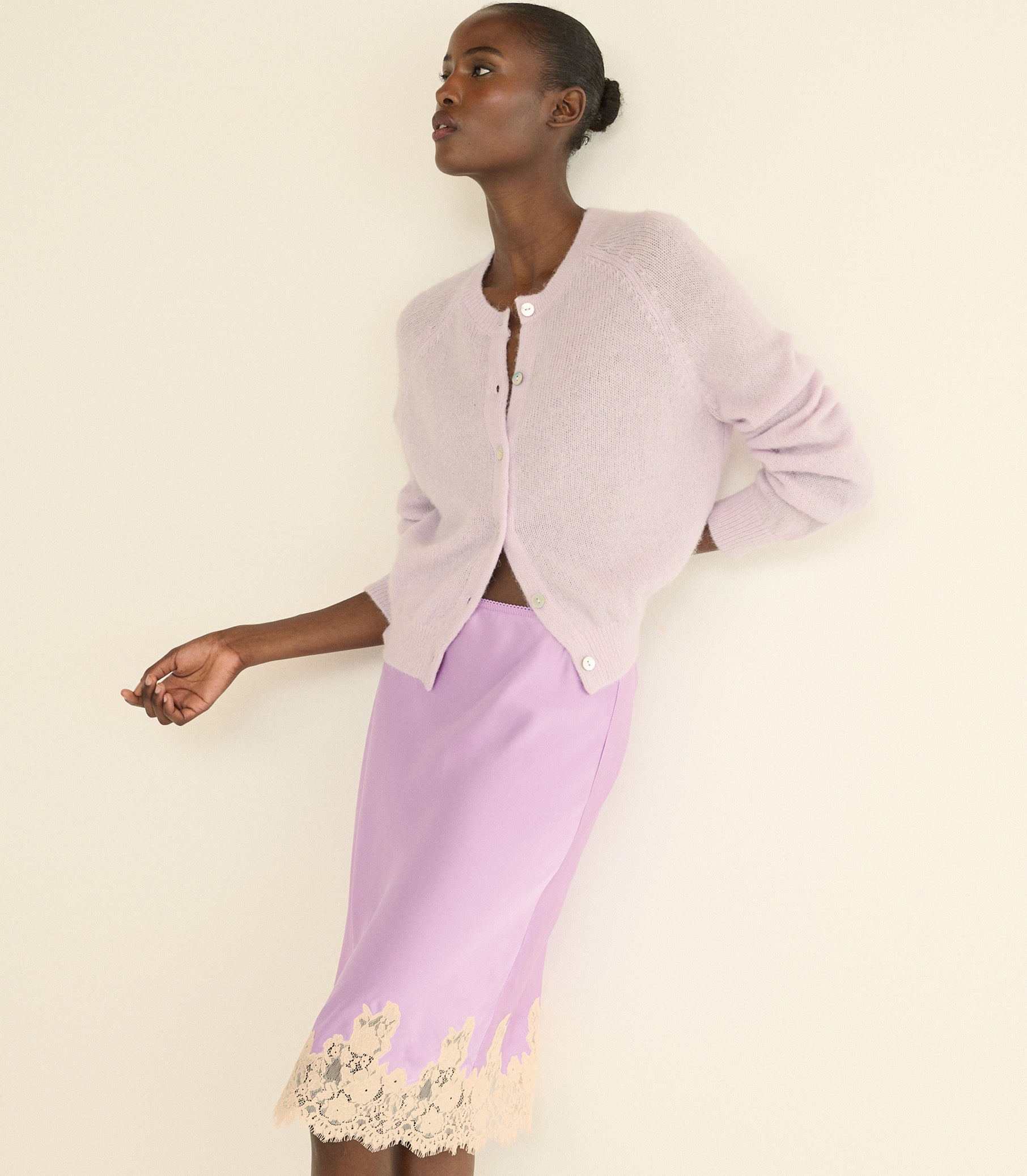 A model wearing a light lavender knit cardigan and a matching lavender silk skirt with a cream lace hem, posed against a solid off-white background.
