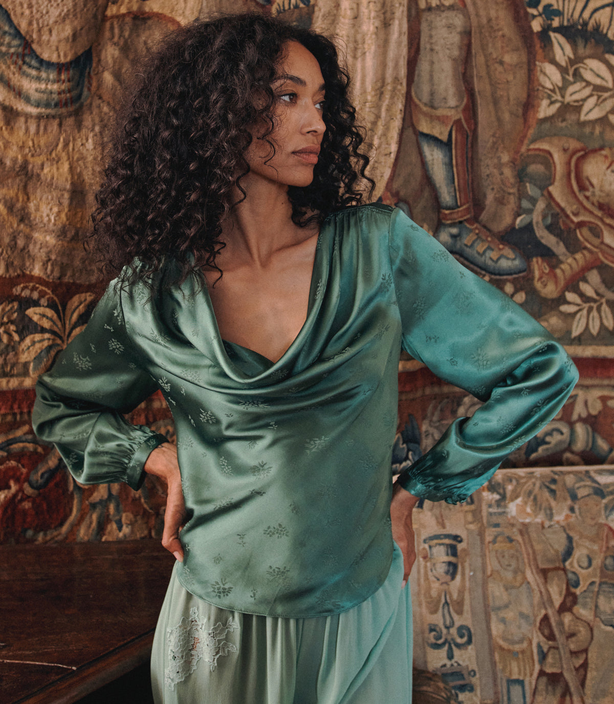 A woman with curly dark hair wearing a sage green satin long-sleeve top and matching skirt, posing in front of a detailed tapestry. 1
