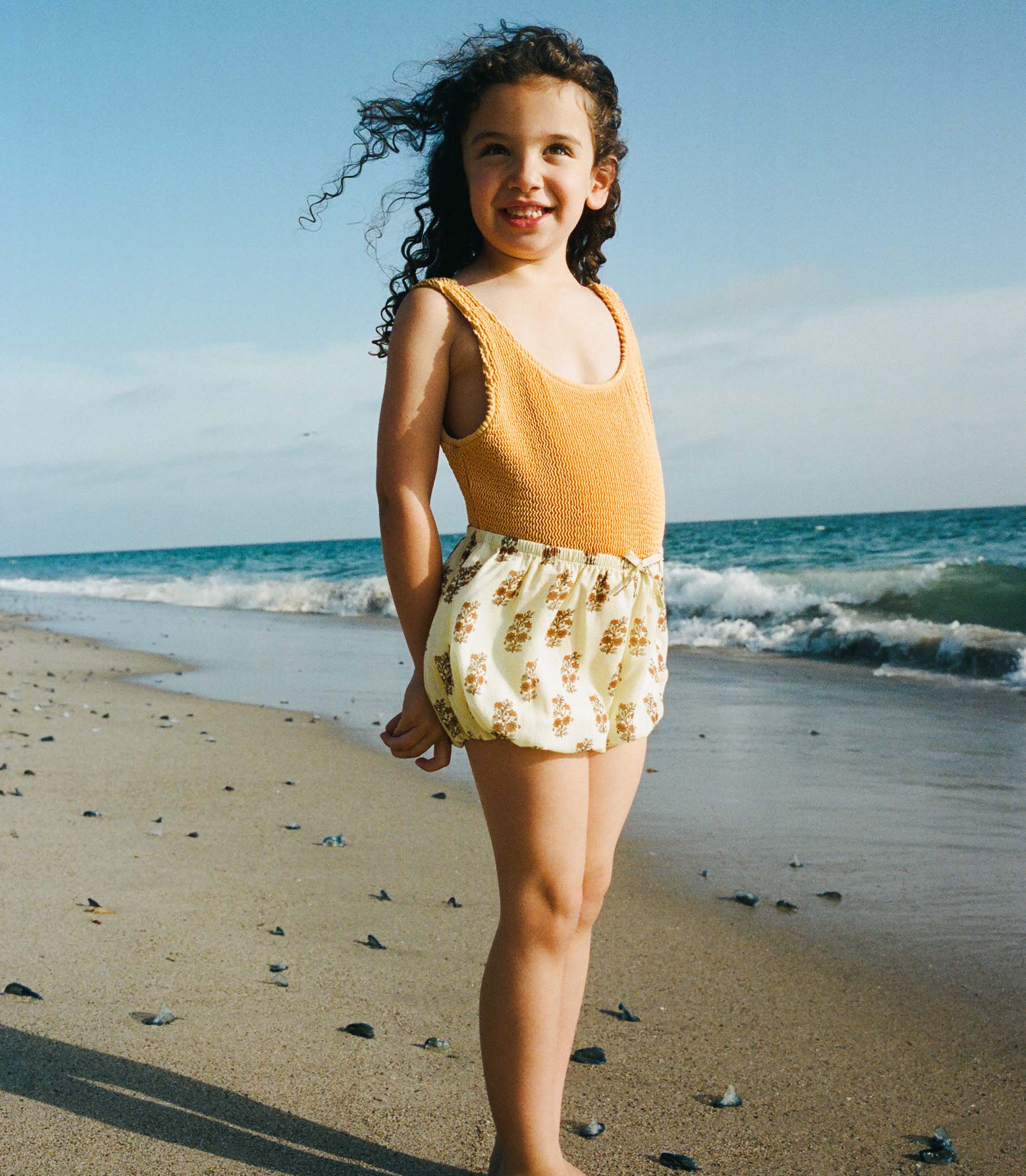 Young girl in a yellow top and floral shorts standing on a beach with ocean waves in the background.