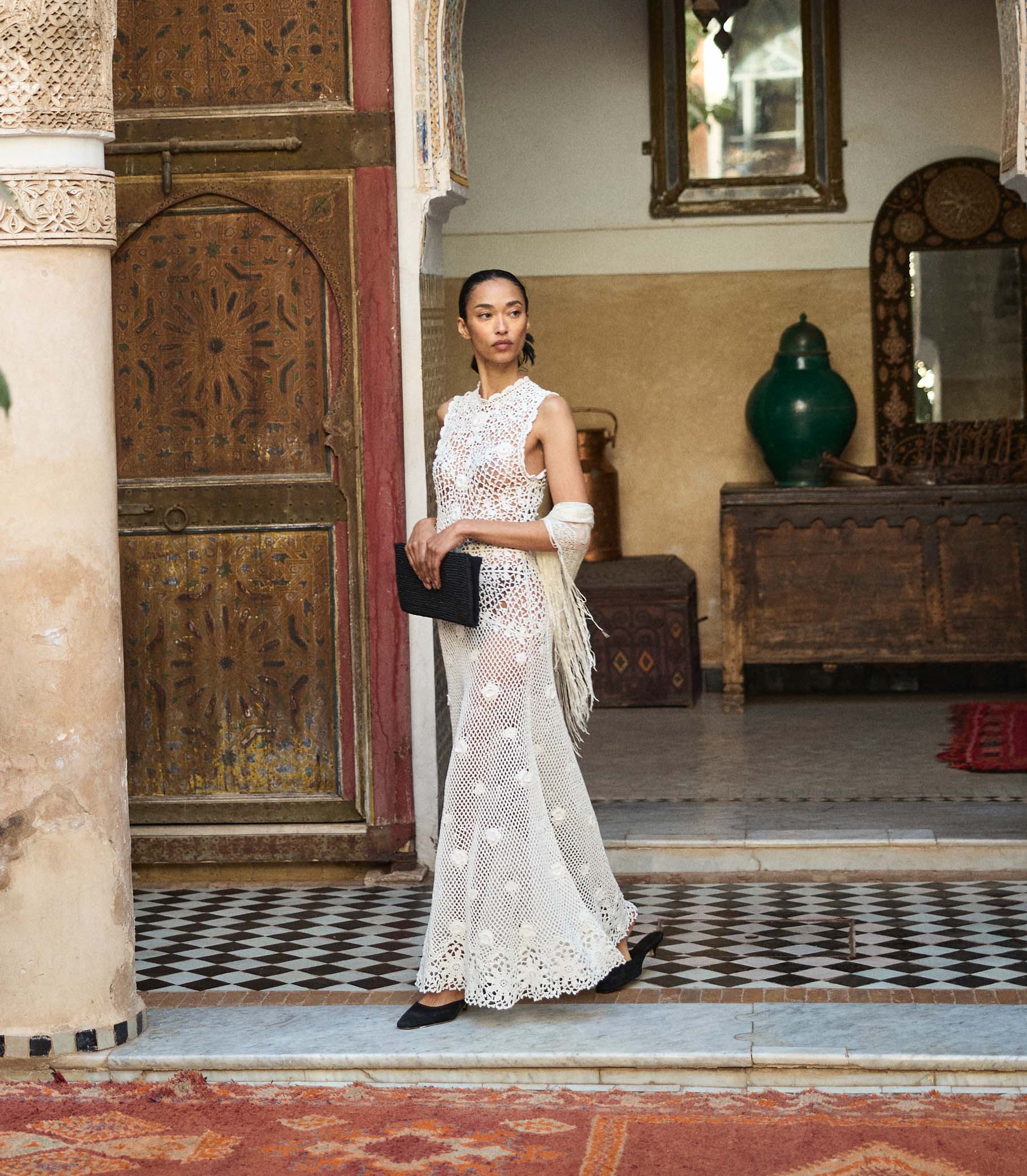 Woman in a white crochet dress standing in an ornate room with decorative elements. 1
