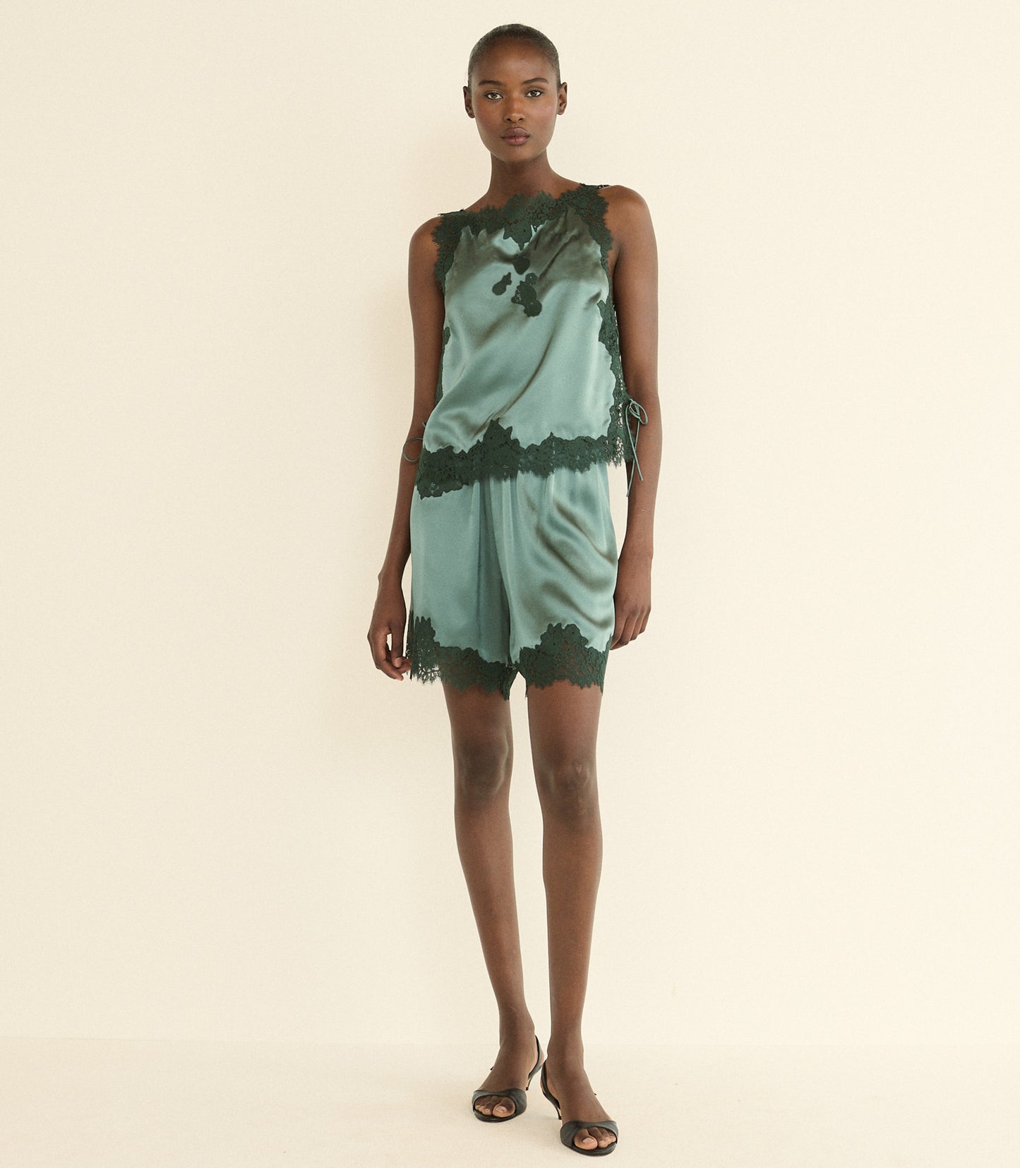 A model stands against a plain cream studio background wearing a sleeveless green satin top with dark green lace trim and a dark green abstract print.