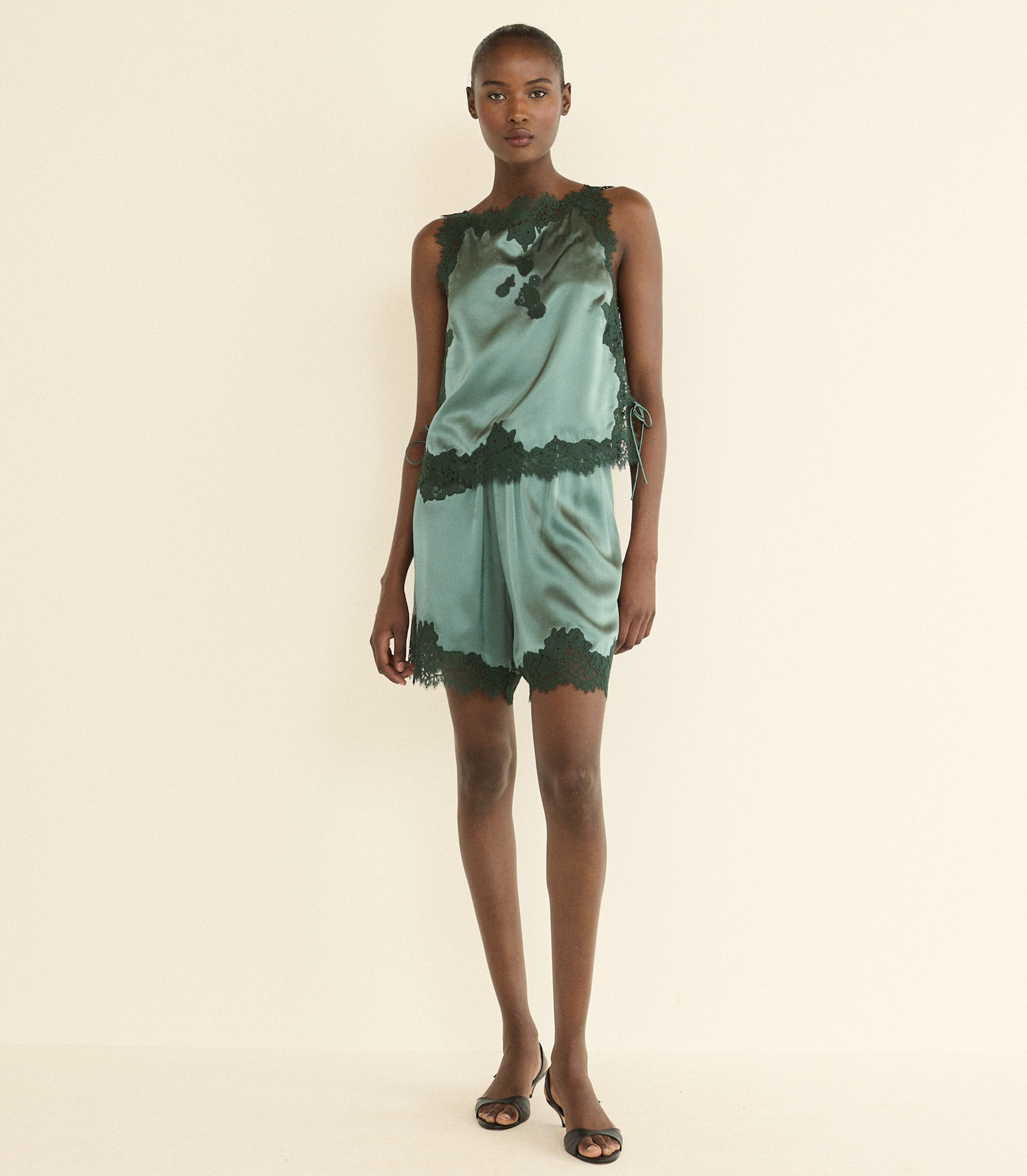A model stands against a plain cream studio background wearing a sleeveless green satin top with dark green lace trim and a dark green abstract print.