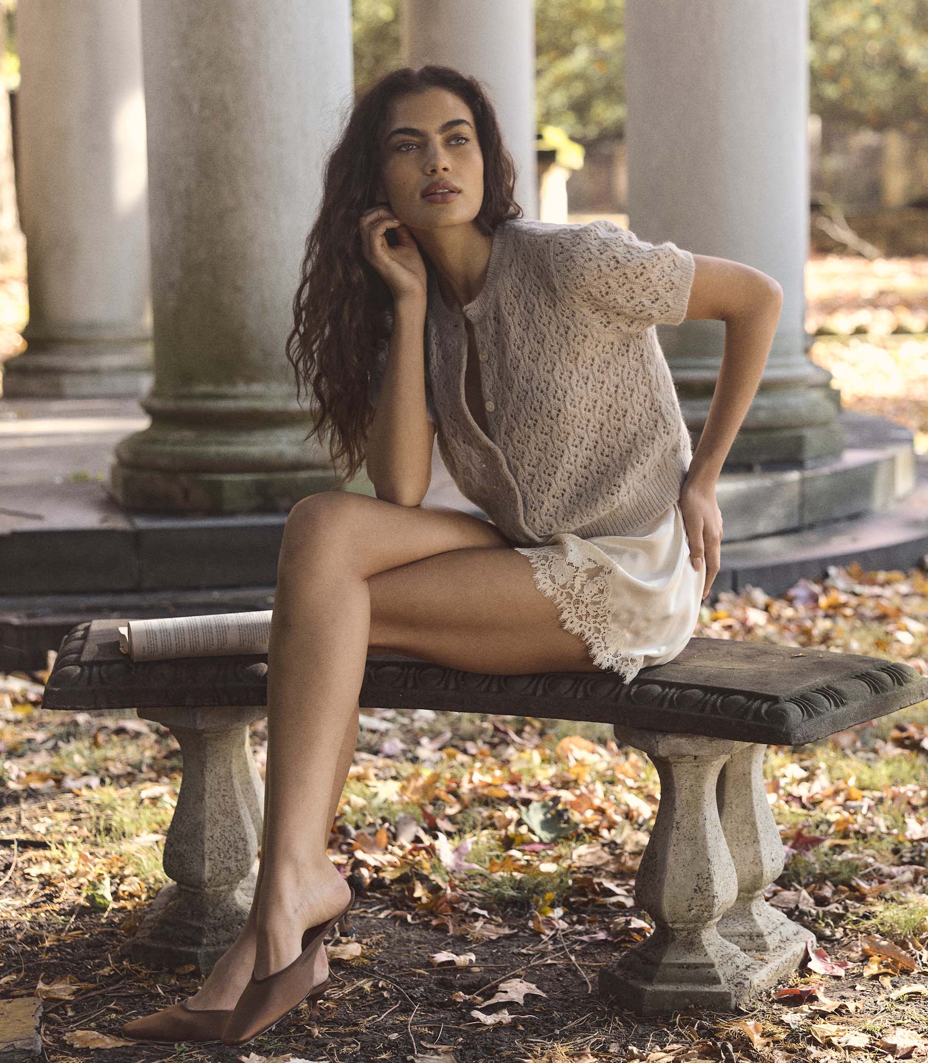 Woman wearing a beige short sleeve cardigan sitting on a stone bench in a park with columns in the background