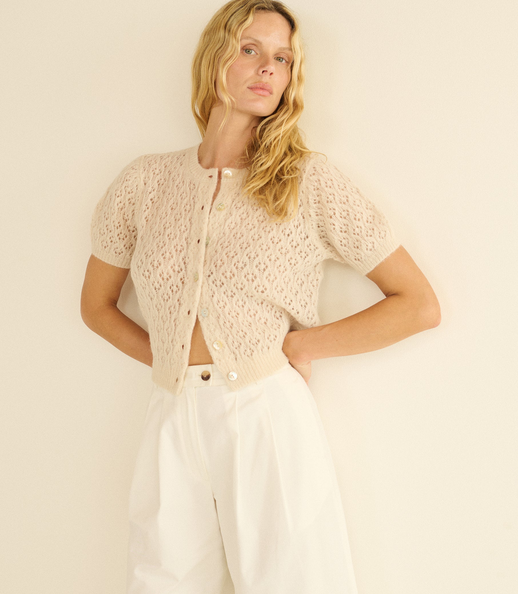 Woman wearing a beige knitted cardigan over a white top and pants on a plain background 1
