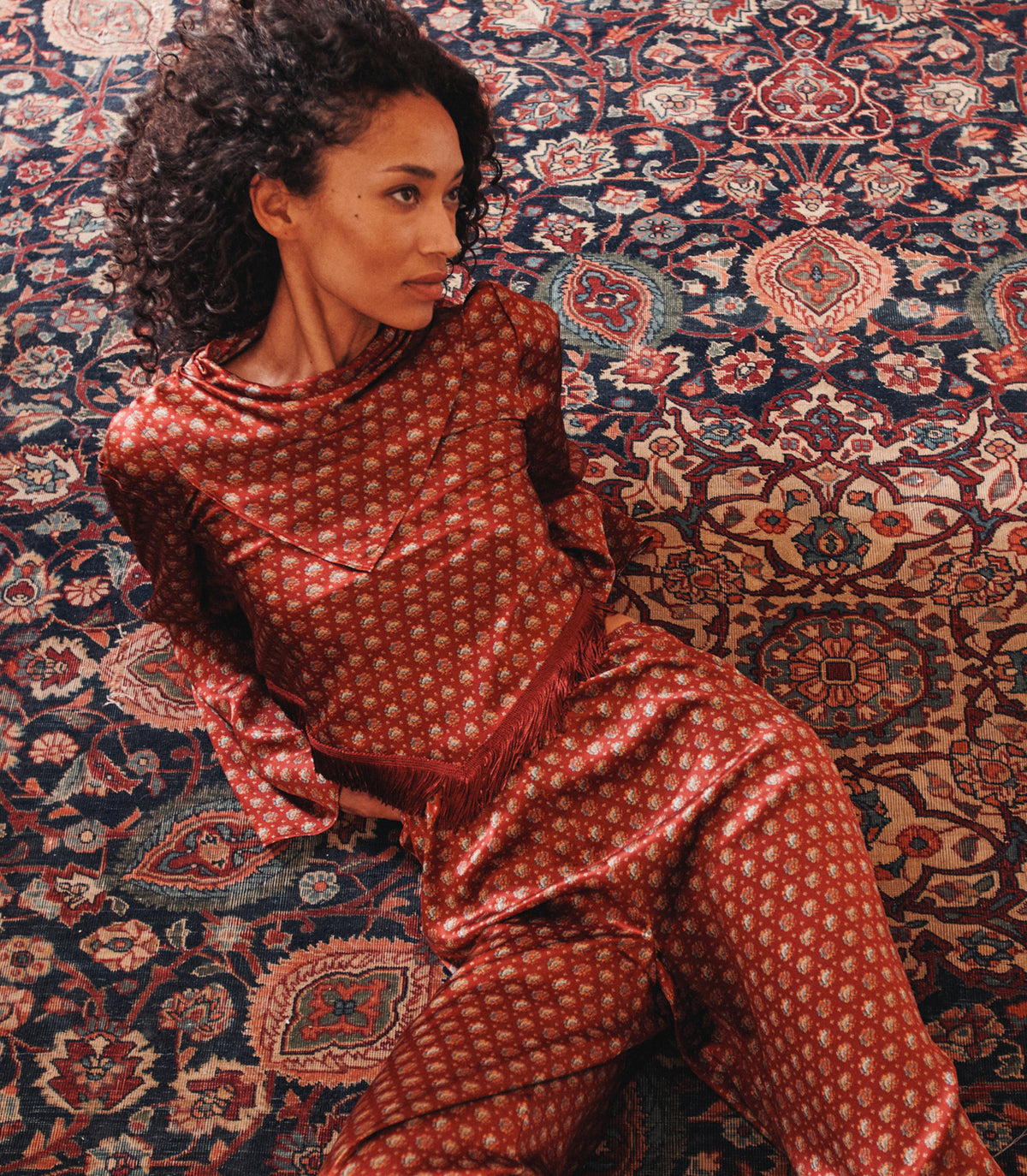 A woman reclining on a detailed oriental rug wearing a matching red and gold patterned pajama set.