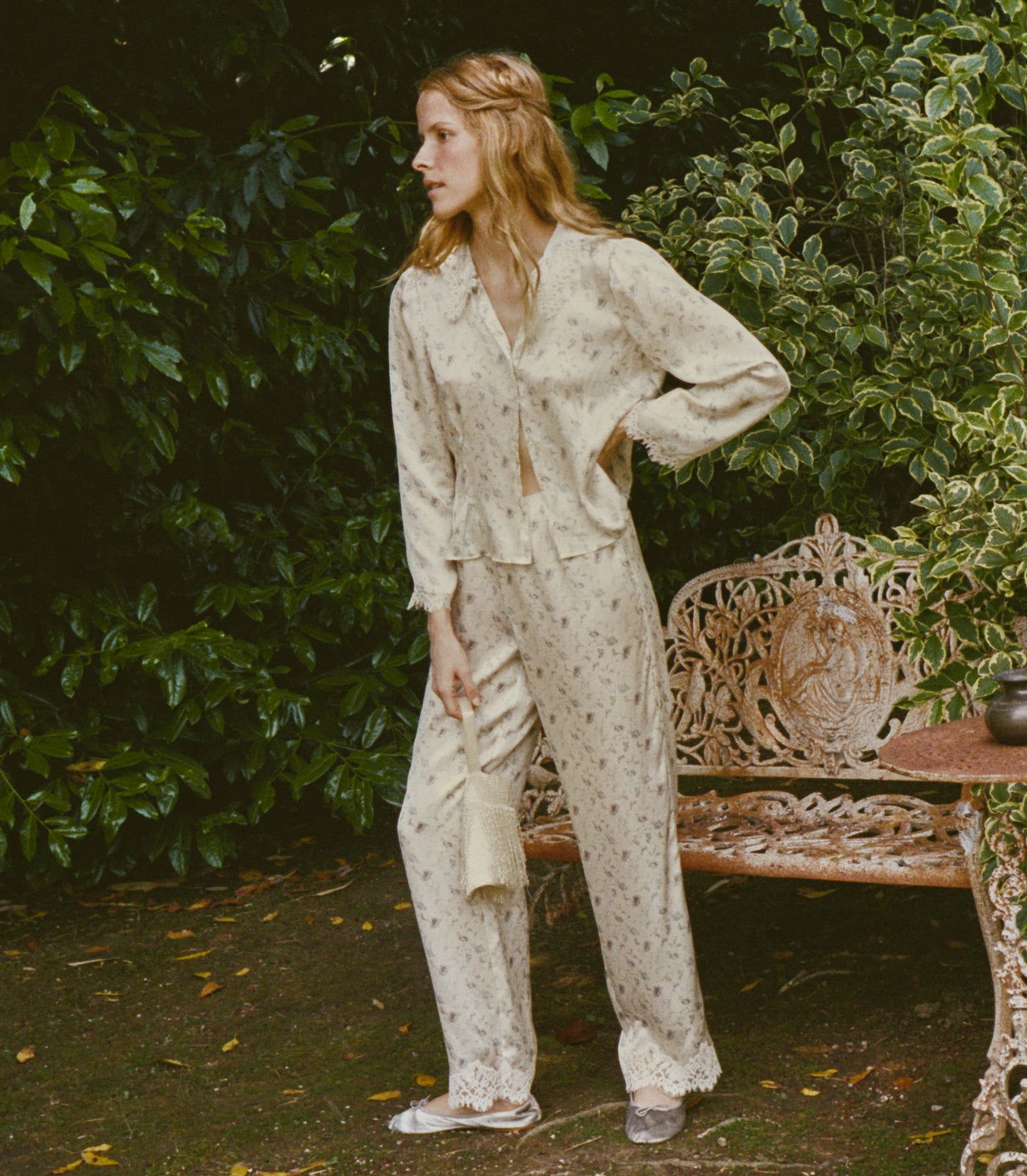 A woman stands outdoors in a garden setting wearing a light cream silk set with a small floral print. Sporting a long-sleeve button-front top and matching wide-leg pants.