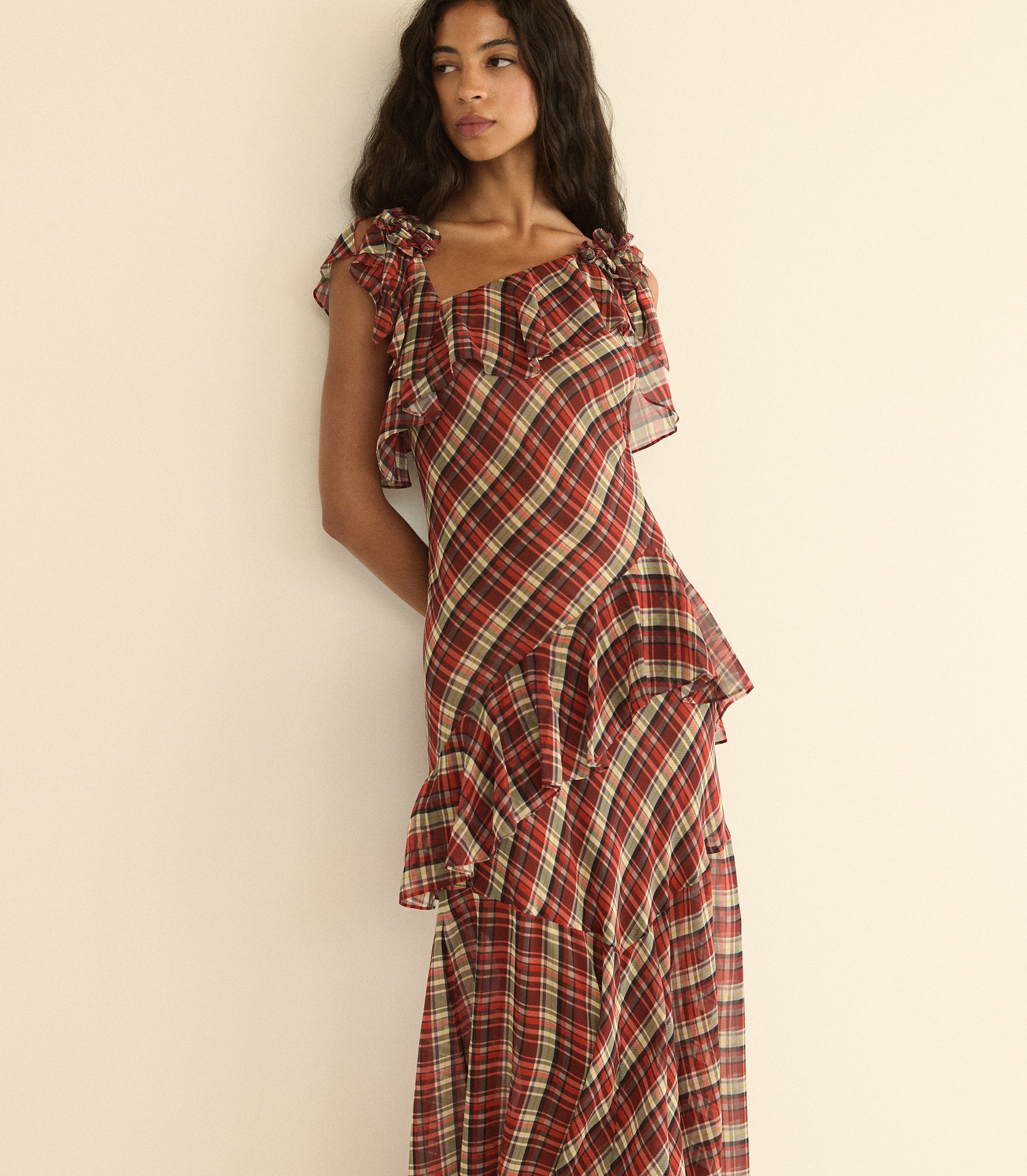 A woman wearing a red and cream plaid ruffled dress, posing against a plain beige wall. 1
