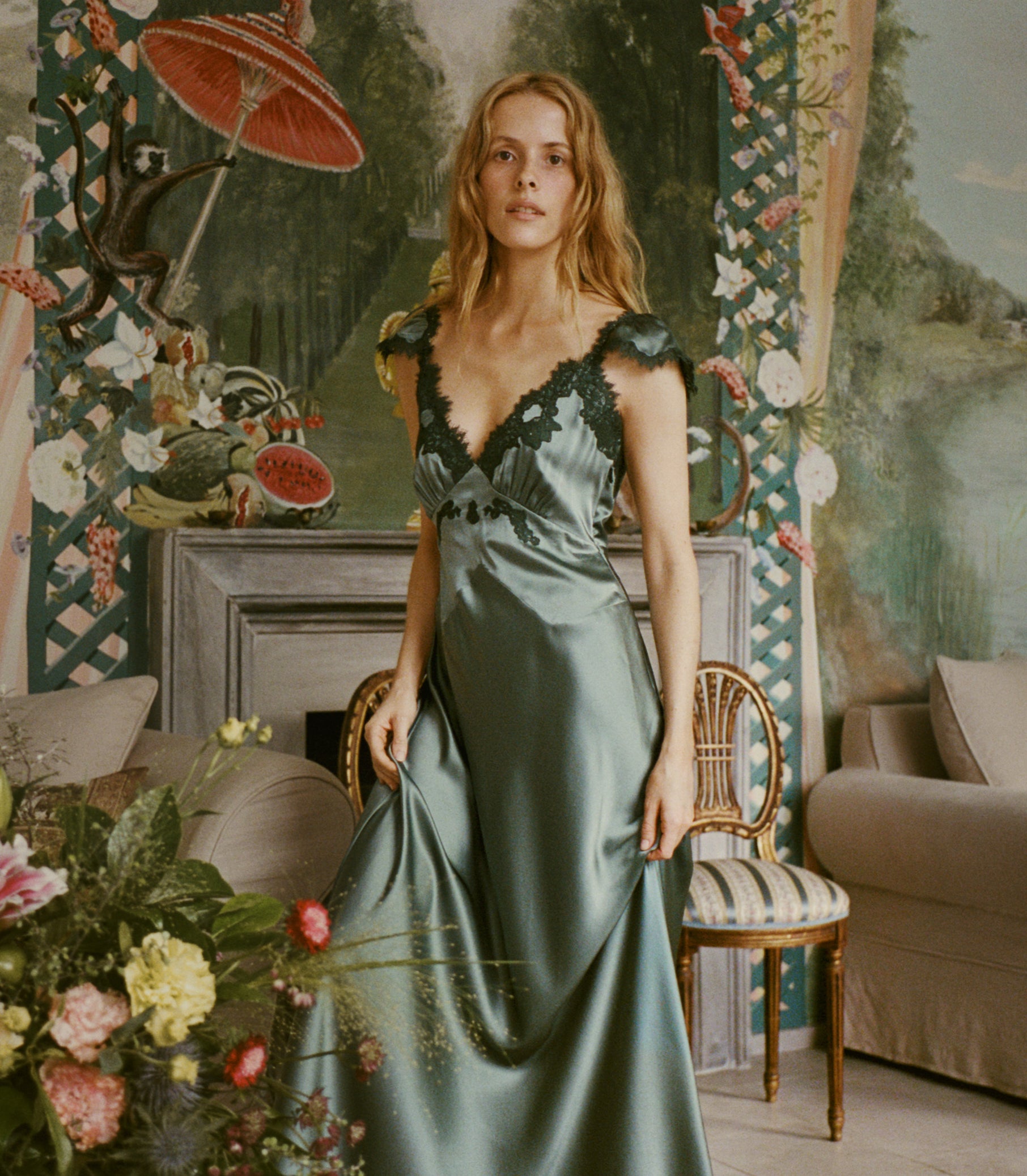 A woman stands in an ornate interior wearing a satin evening gown. The dress is a muted sage green with dark lace trim at the neckline and shoulders.