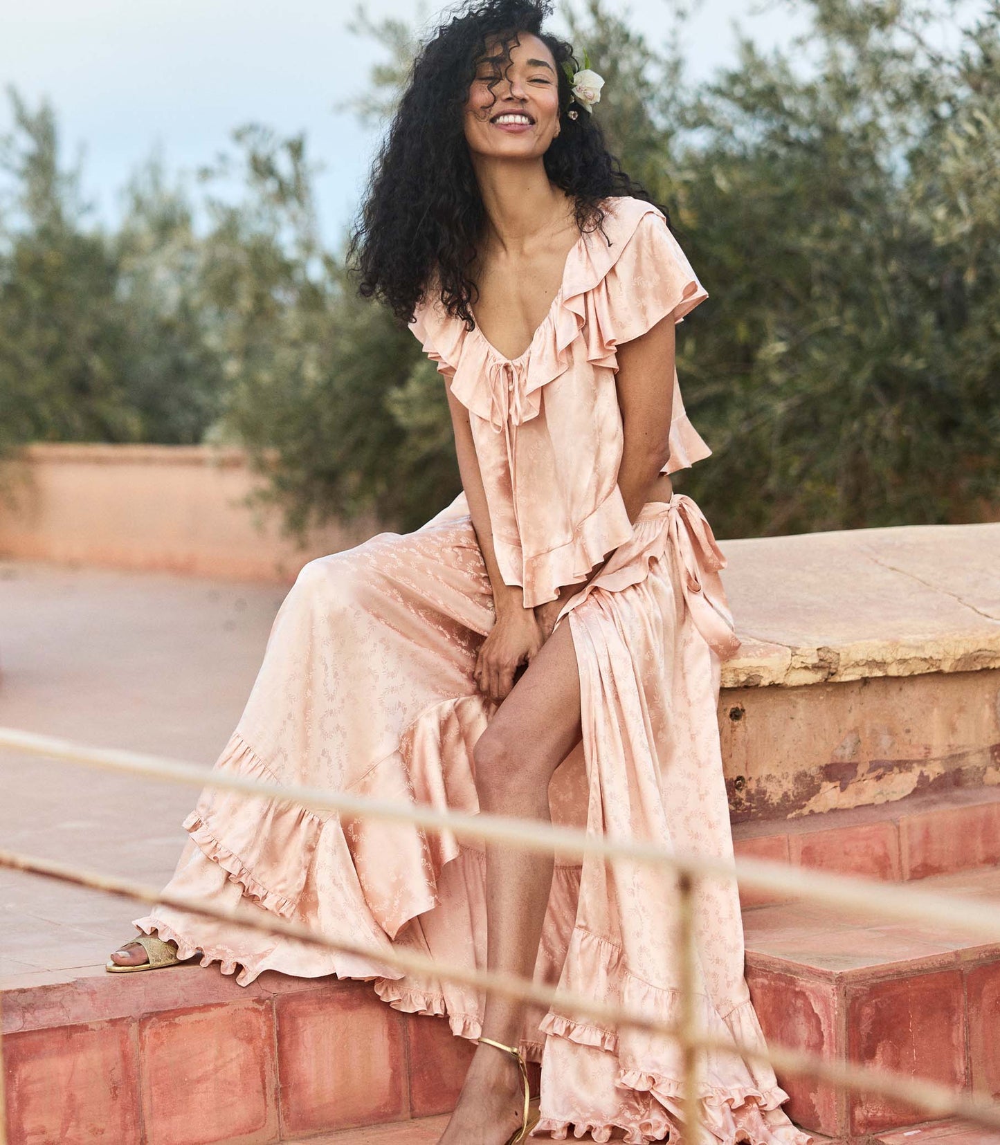 Woman in a flowing peach matching set sitting outdoors with greenery in the background