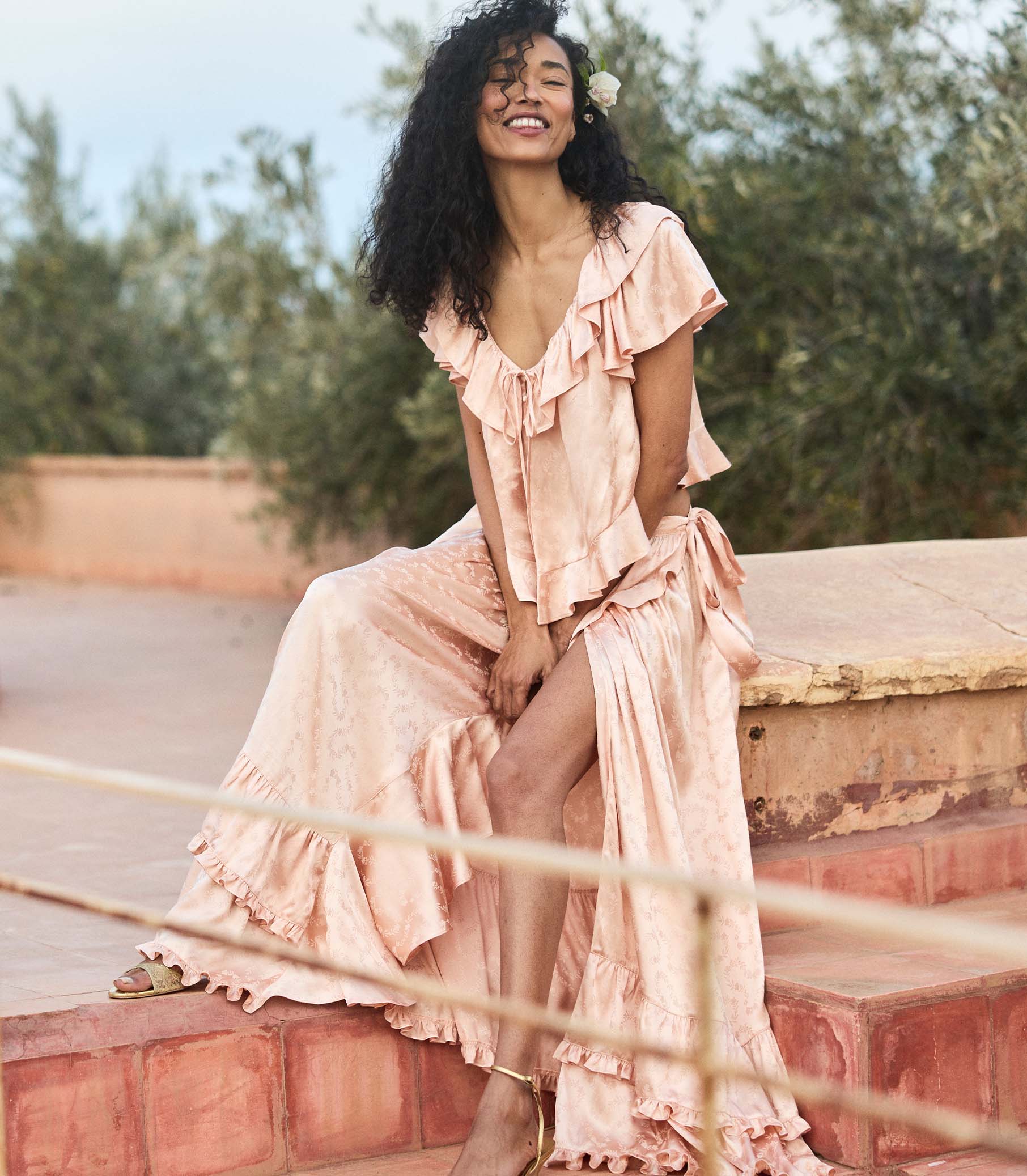 Woman in a flowing peach matching set sitting outdoors with greenery in the background