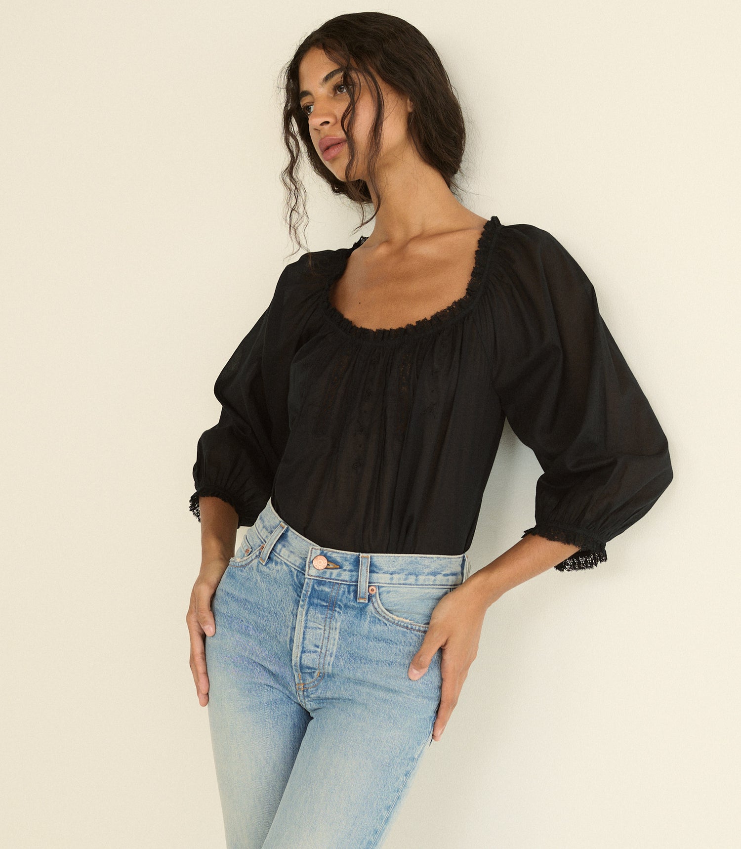 A woman wearing a black puff-sleeve blouse with a square neckline and scalloped trim, paired with light blue jeans, posing against a plain off-white background.