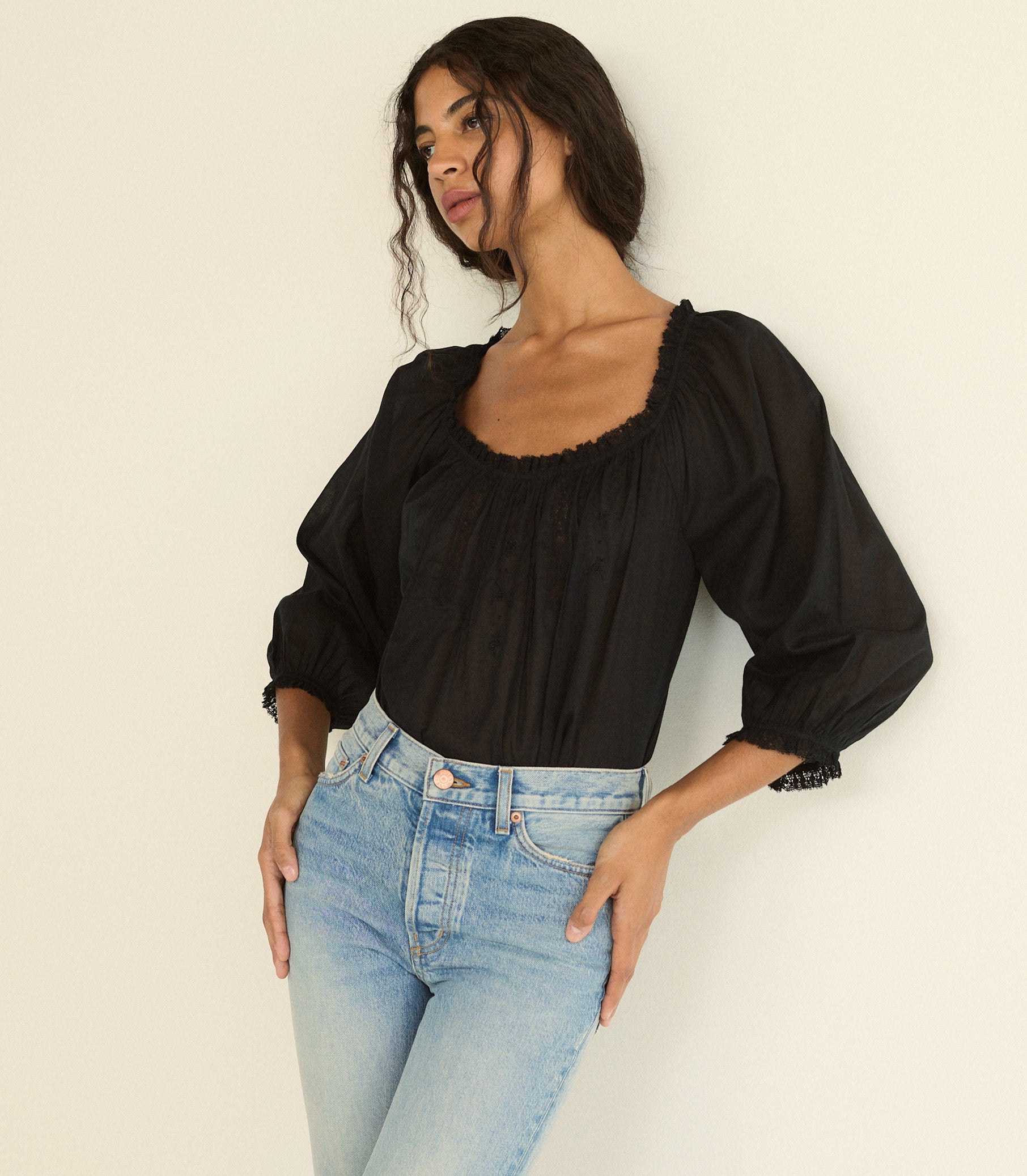 A woman wearing a black puff-sleeve blouse with a square neckline and scalloped trim, paired with light blue jeans, posing against a plain off-white background. 1
