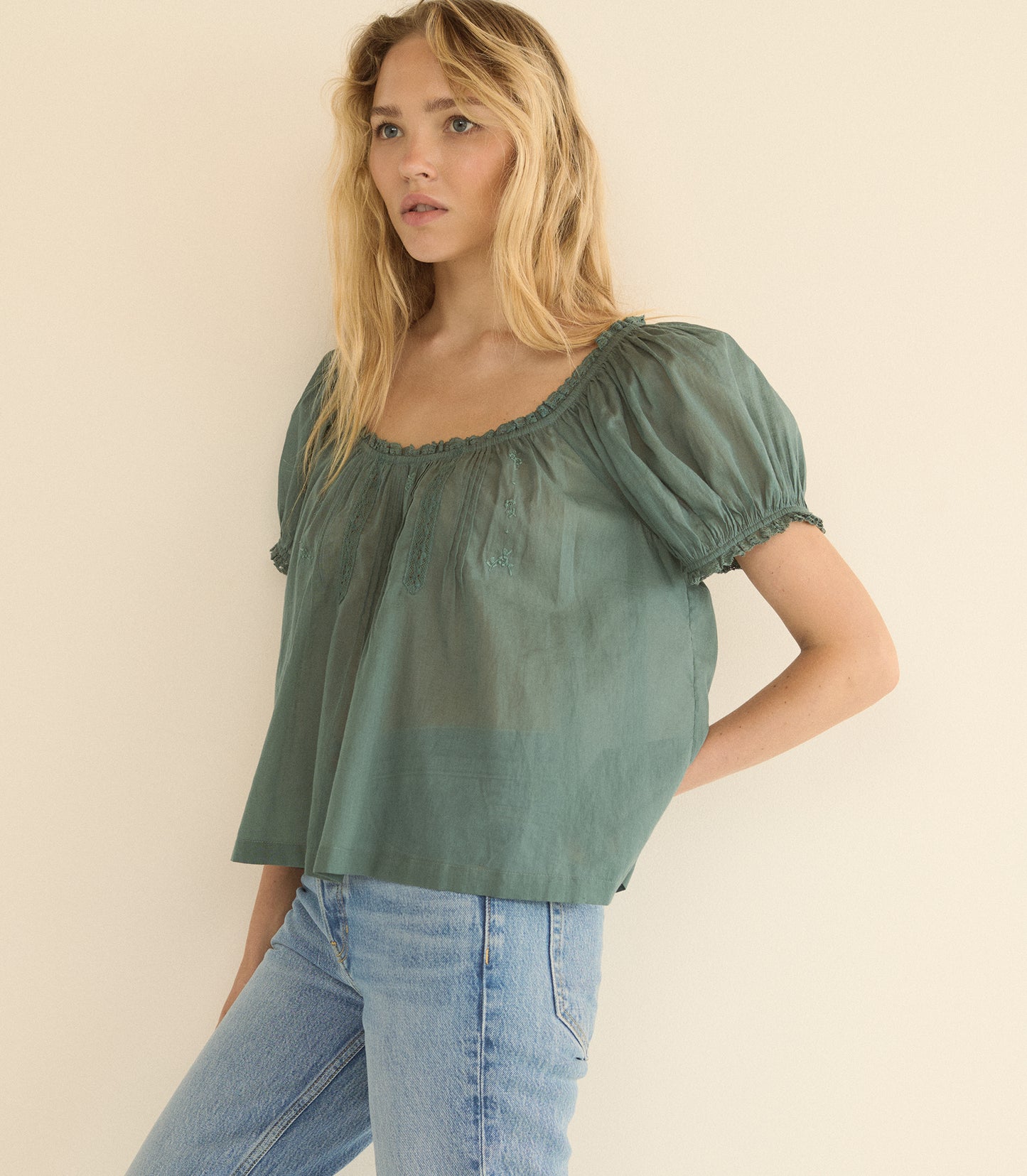 A woman wearing a sage green puff-sleeve top and light blue jeans, standing against a plain beige background.