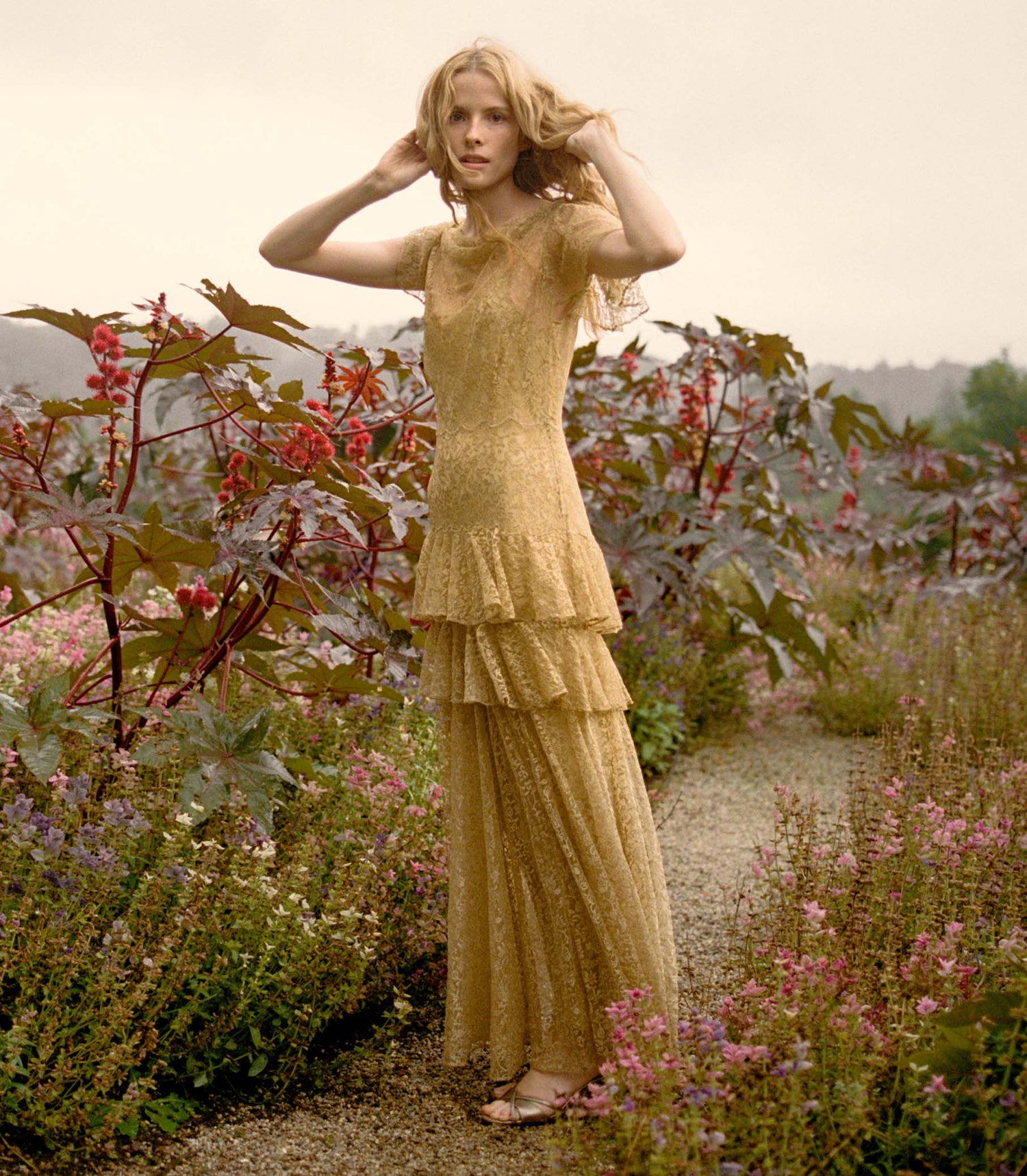 Woman in a gold lace dress standing in a garden with flowers and plants.
