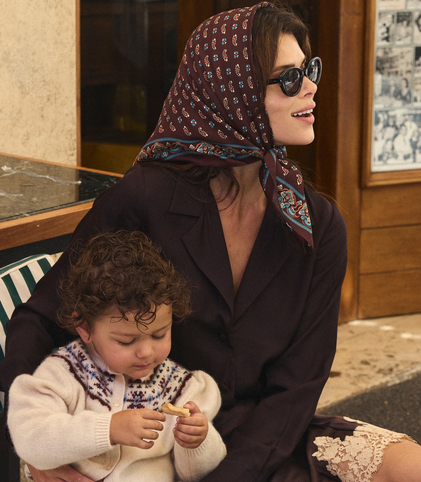 A woman wearing a dark brown patterned headscarf and sunglasses sits outdoors holding a young child. The woman's outfit includes a dark brown button up shirt and a lace-trimmed skirt, while the child wears a cream knit sweater with a patterned collar.