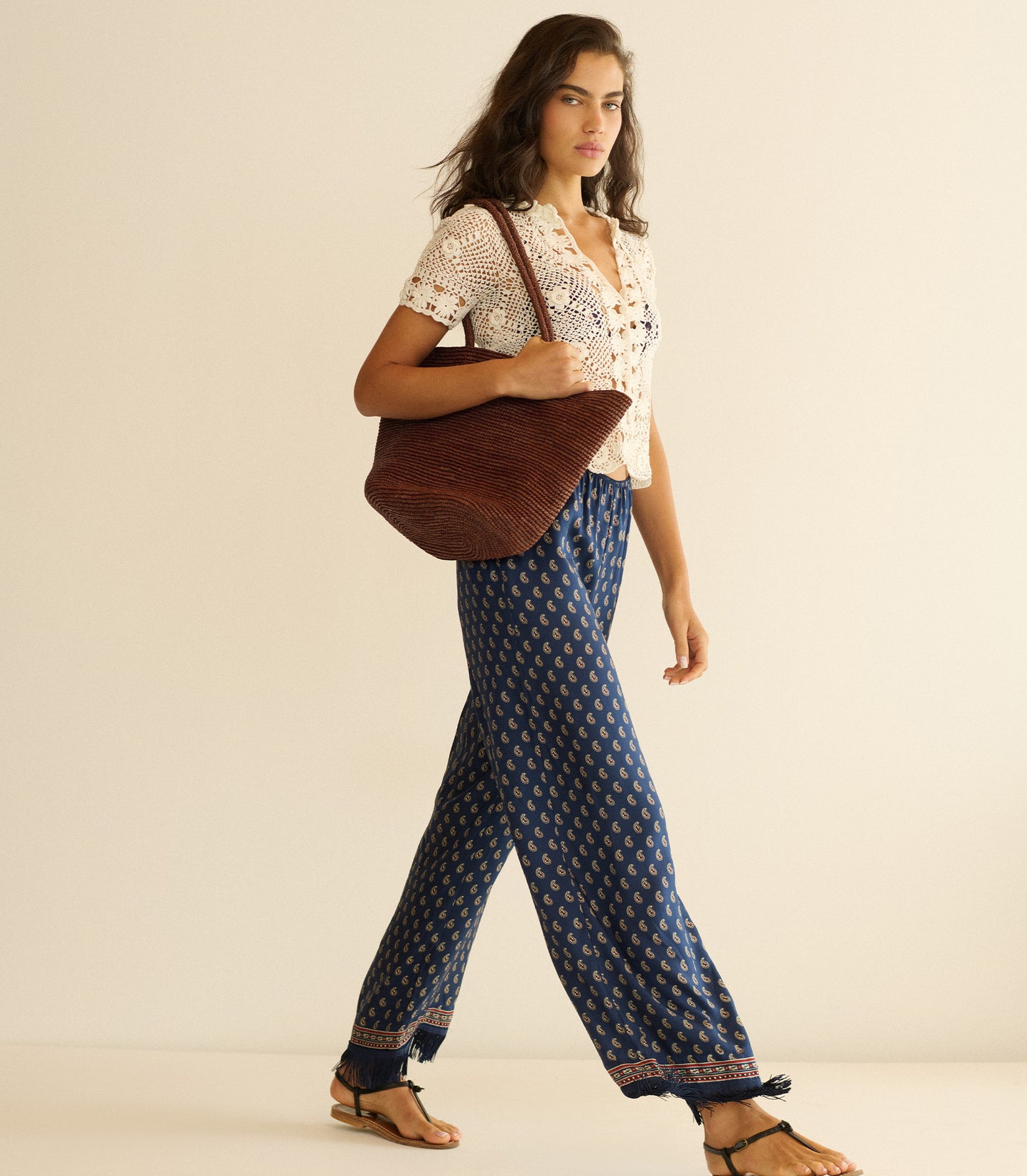 Woman wearing a white blouse and blue patterned pants, holding a brown tote bag on a beige background