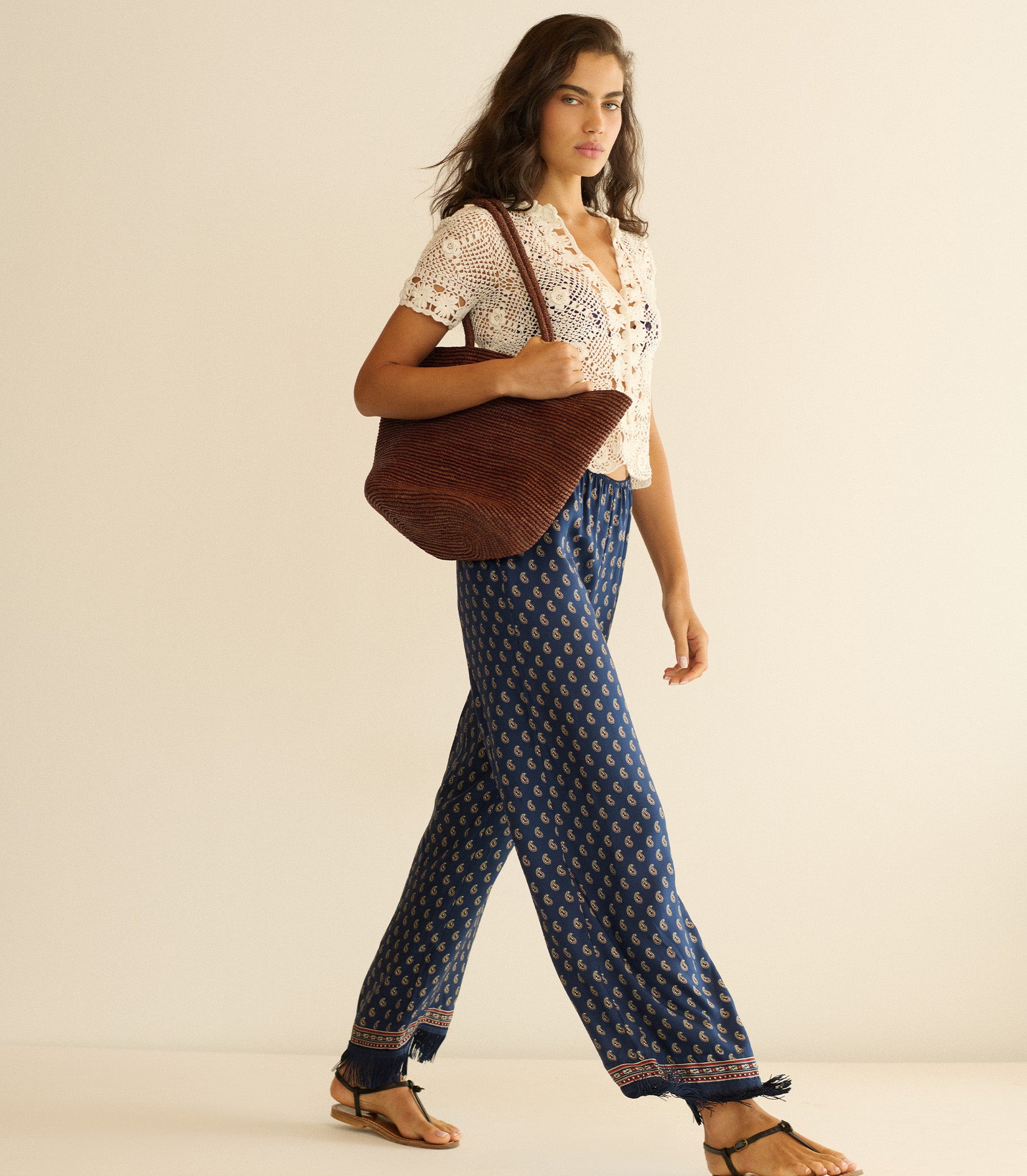 Woman wearing a white blouse and blue patterned pants, holding a brown tote bag on a beige background 1
