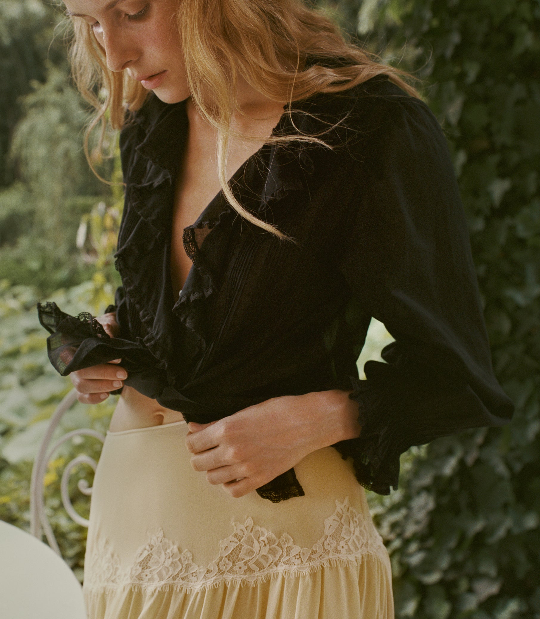 A woman outdoors wearing a black ruffled blouse and a cream lace-trimmed skirt, with green foliage in the background. 1
