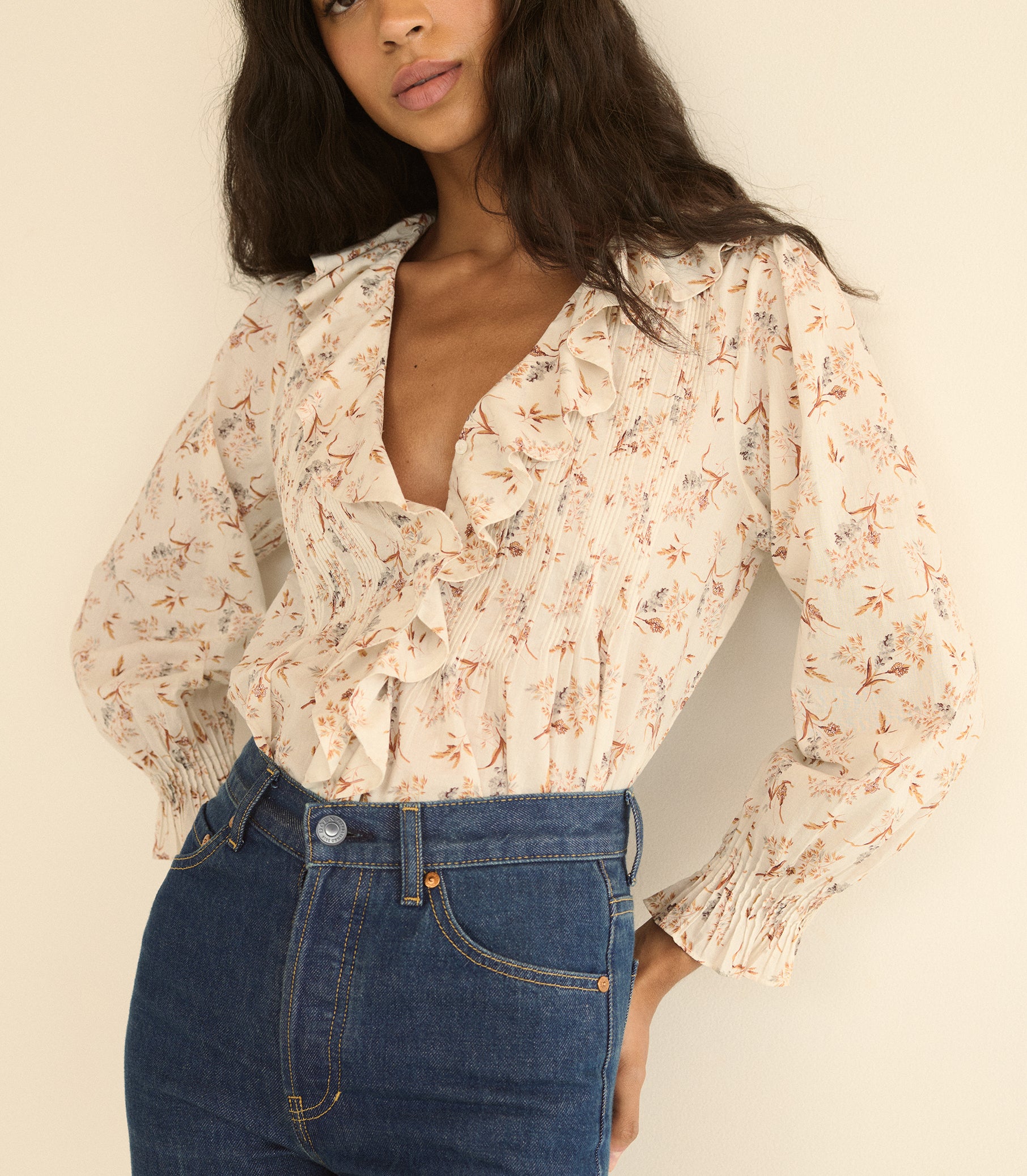 A woman wearing a cream-colored long-sleeve blouse with a delicate orange and blue floral print, paired with high-waisted blue jeans, against a plain beige background. 1
