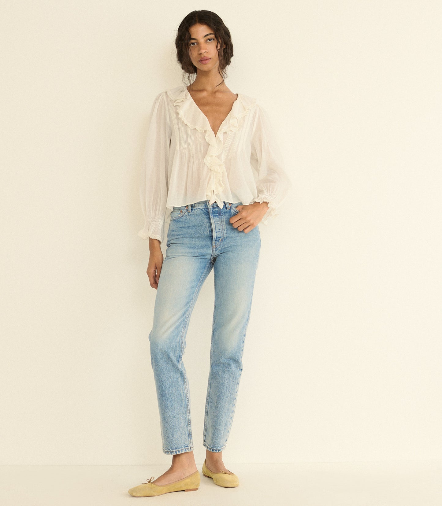 A woman modeling a white ruffled long-sleeve blouse and light-wash blue jeans against a plain off-white studio background.