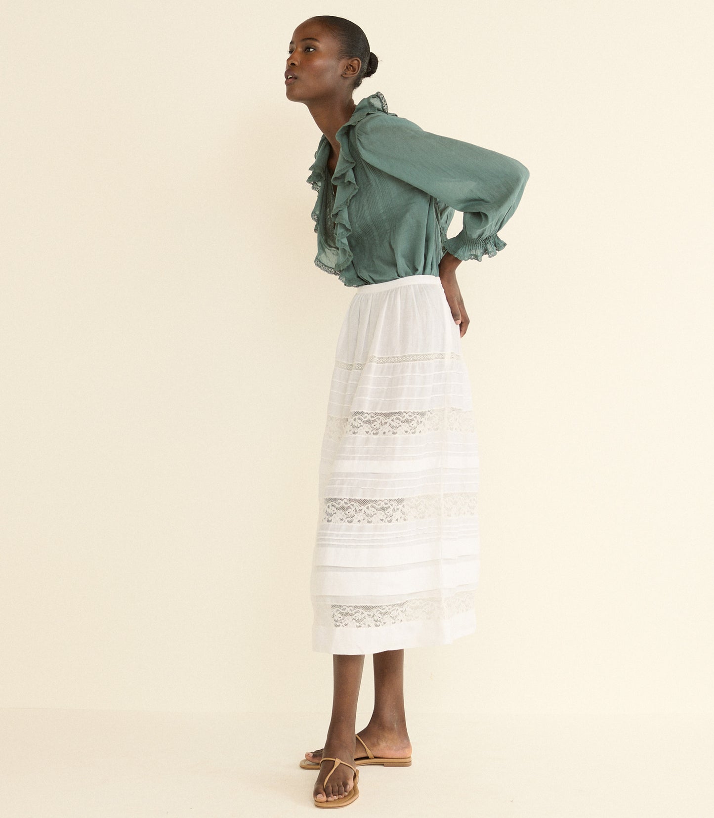 A model is shown from the side wearing a sage green ruffled blouse and a white tiered skirt with lace details, paired with brown flat sandals, against a solid cream background.