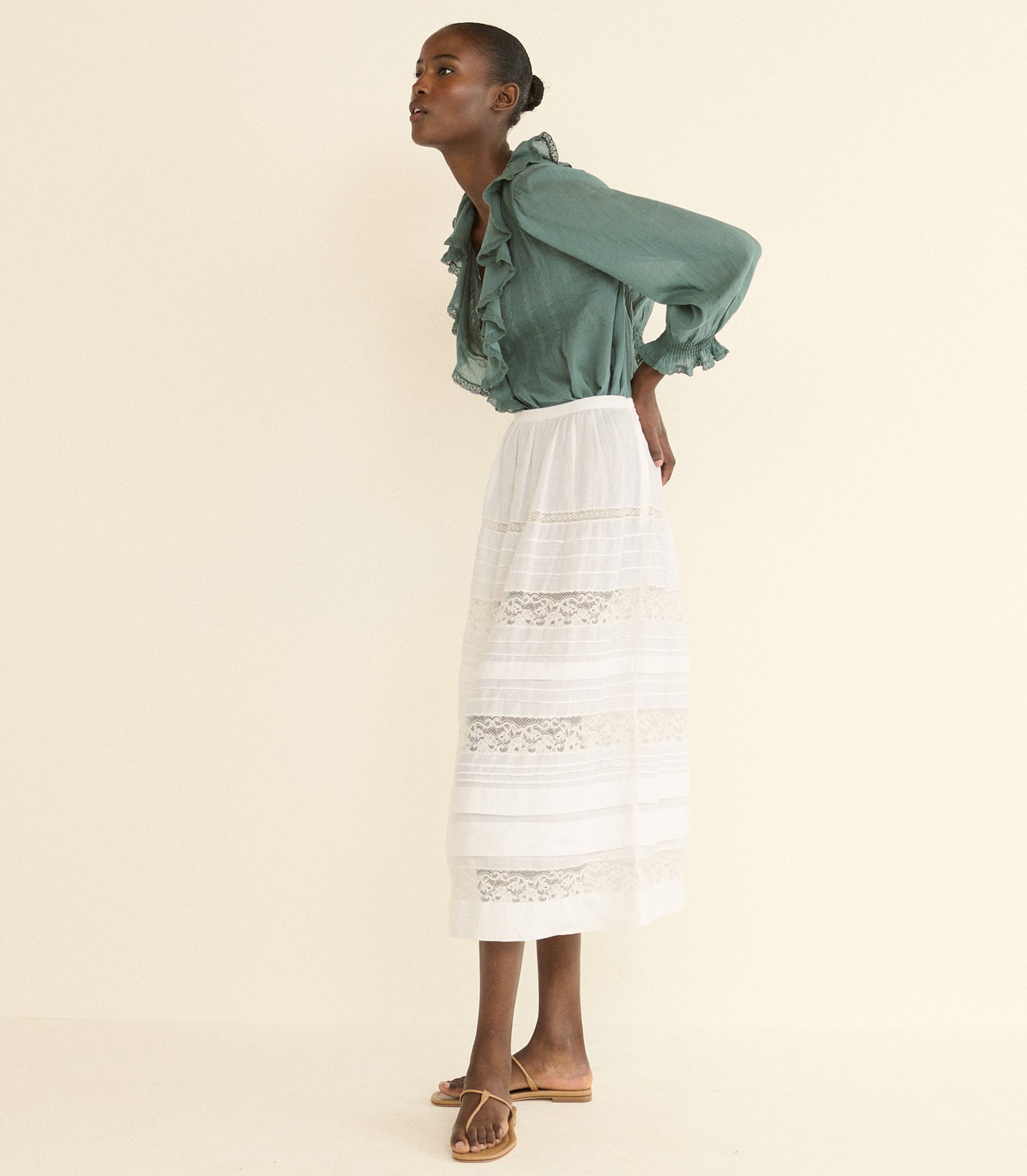 A model is shown from the side wearing a sage green ruffled blouse and a white tiered skirt with lace details, paired with brown flat sandals, against a solid cream background.