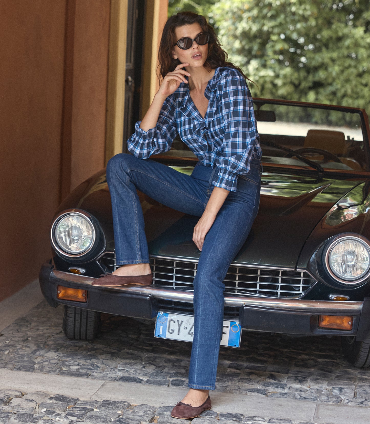 A woman wearing a blue and black plaid button-down shirt, dark blue jeans, and brown flat shoes sits on the front bumper of a vintage black car in an outdoor setting.