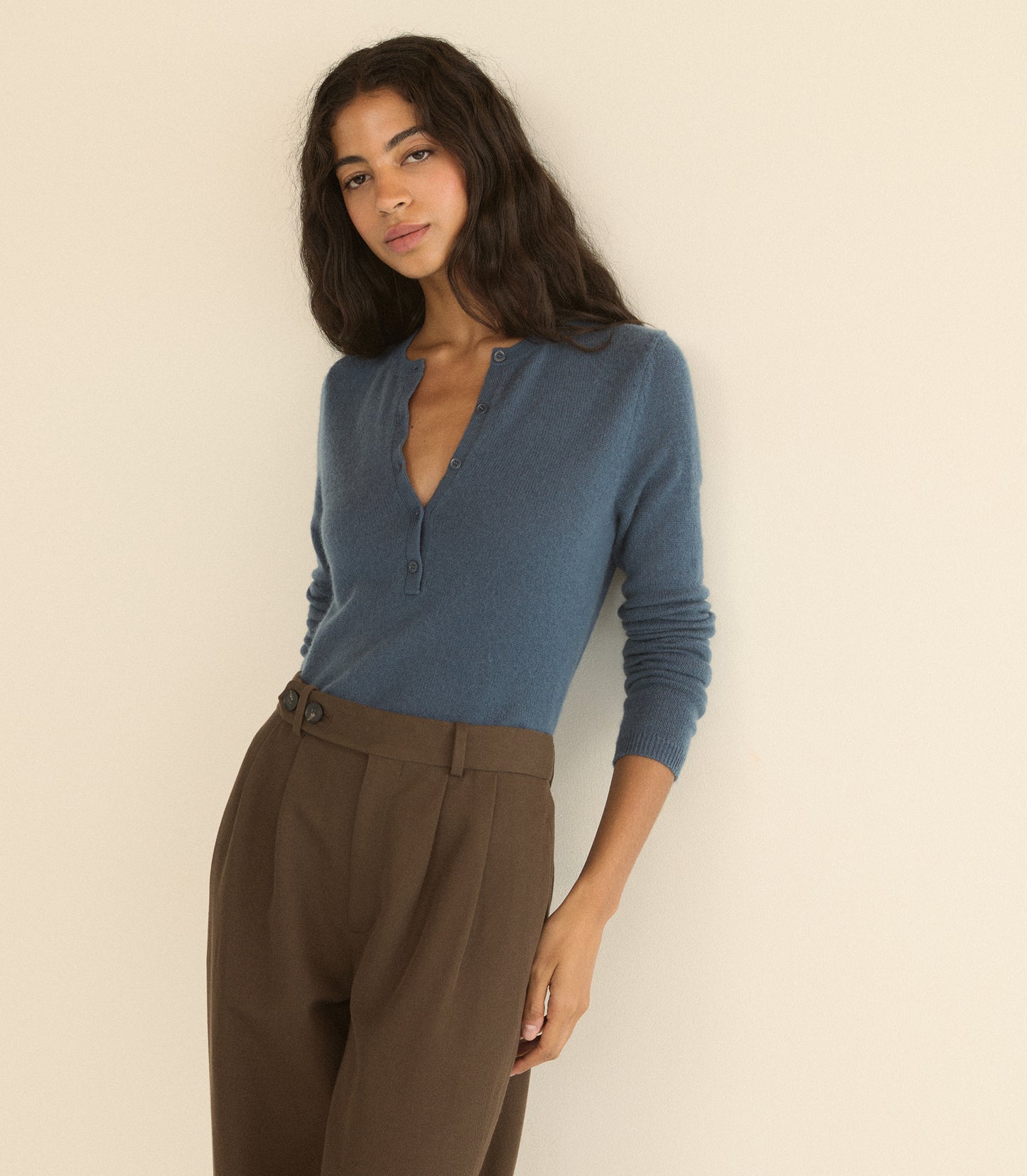 A woman wearing a dusty blue long-sleeve henley top and brown trousers, posing against a plain beige wall.