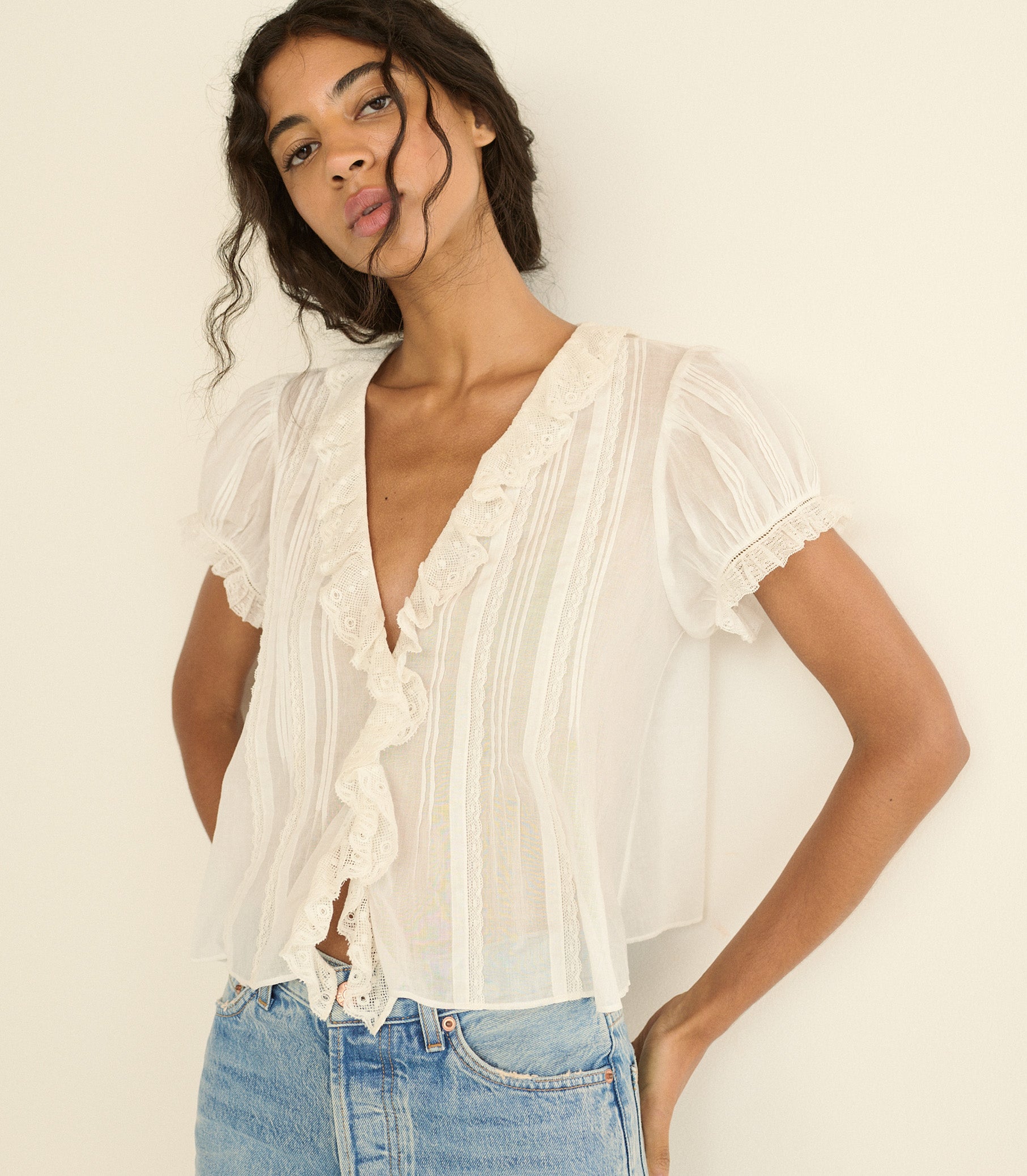 A woman wearing a sheer white short-sleeve blouse with ruffle trim and vertical pleats, paired with light blue jeans, posing against a plain off-white background. 1
