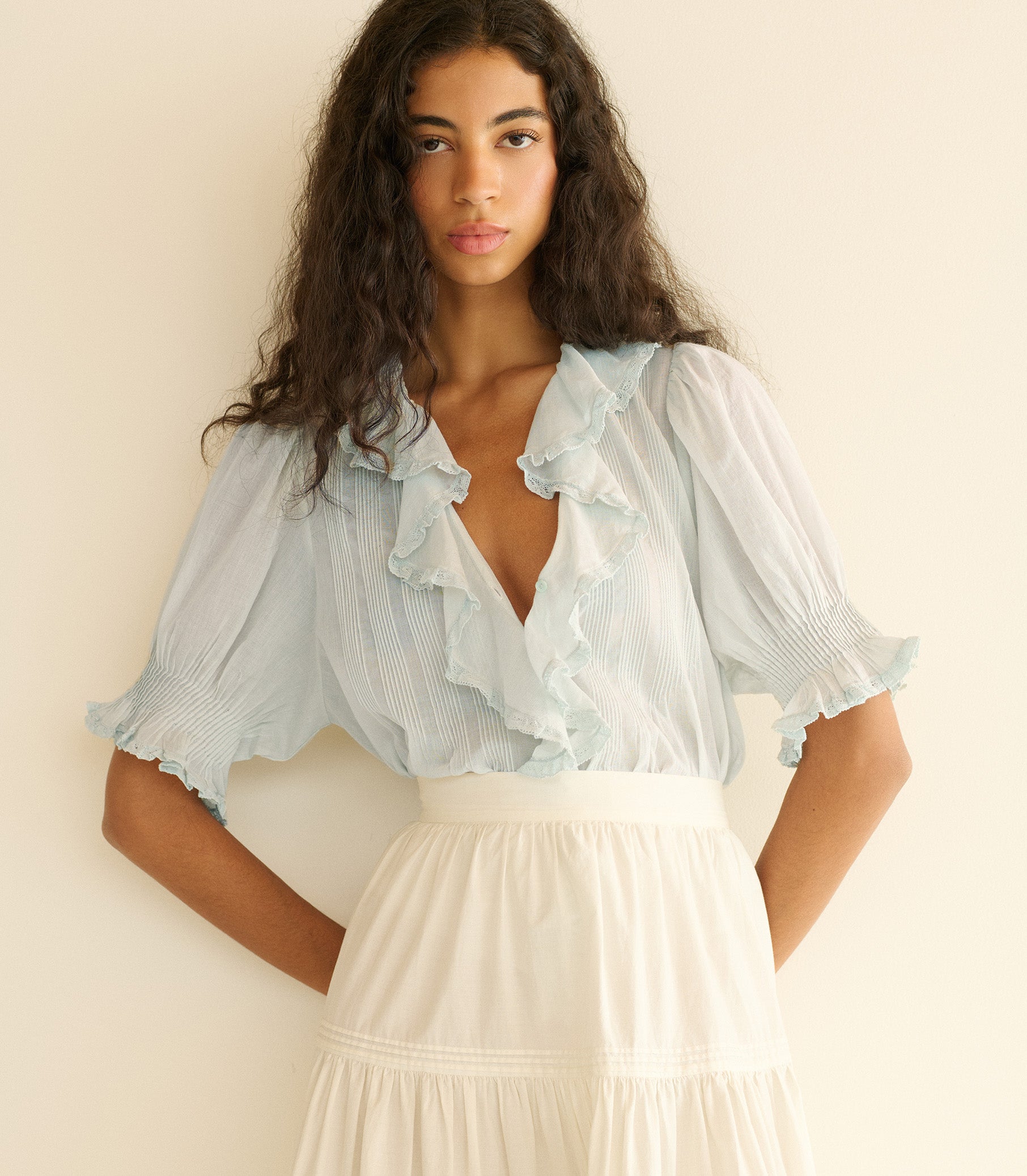 Woman wearing a light blue blouse with ruffles and a white skirt against a beige background 1
