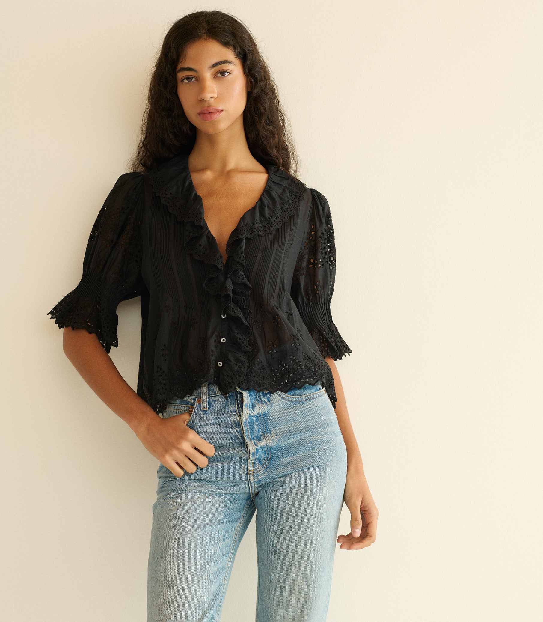 Woman wearing a black blouse with ruffled details and light blue jeans against a beige wall. 1
