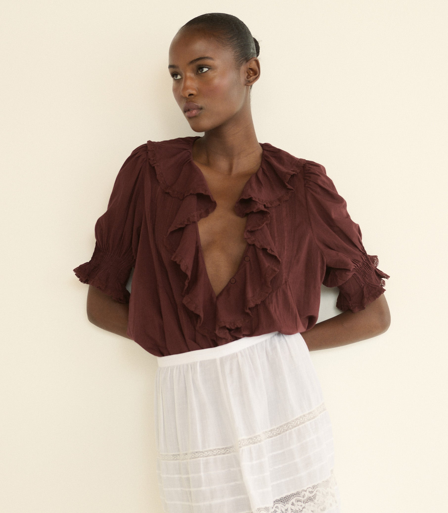 A model wearing a deep burgundy ruffled blouse with puff sleeves and a white skirt, posing against a plain cream-colored wall.