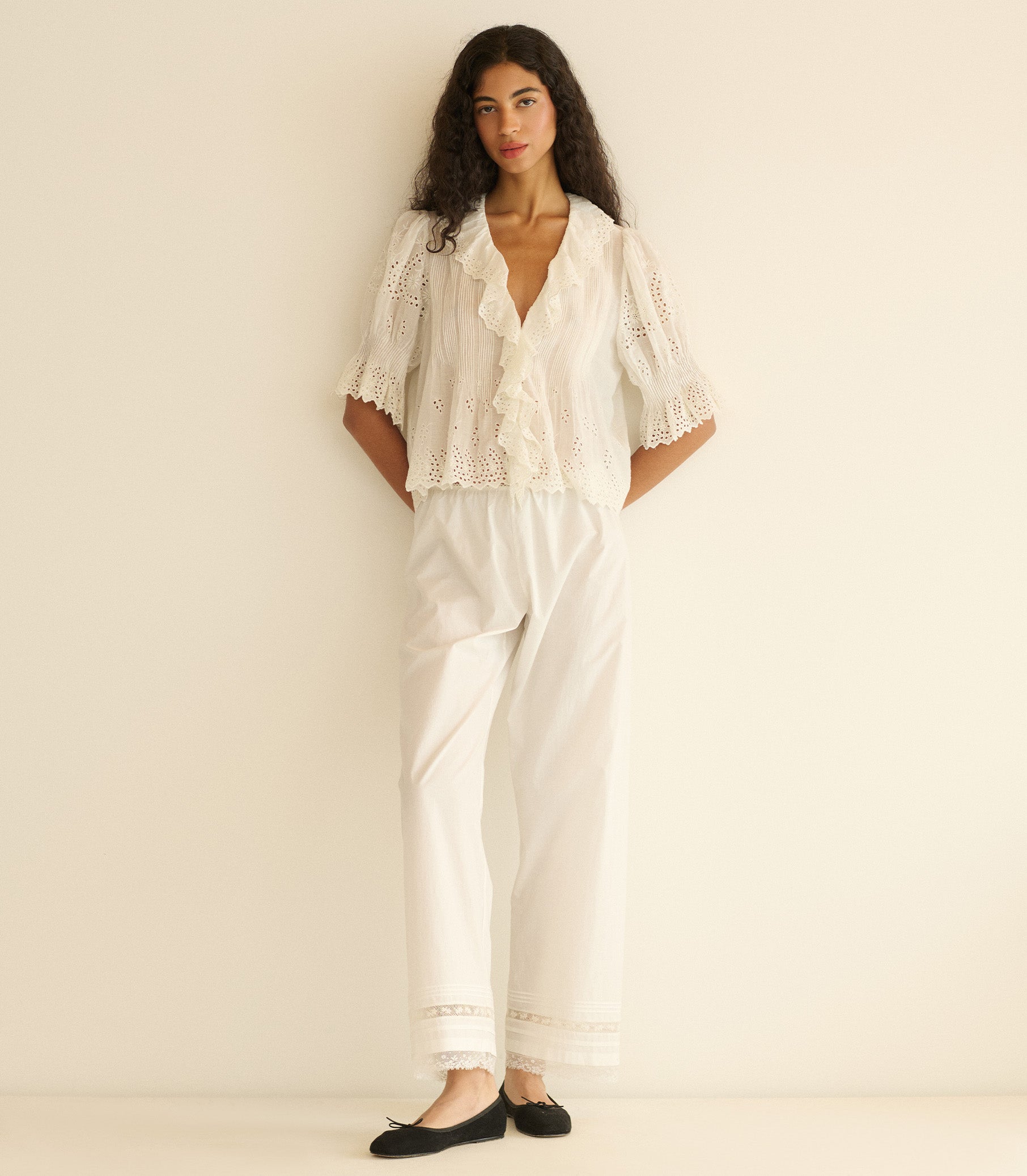 Woman wearing a white eyelet blouse and white pants with lace details on a plain background 4
