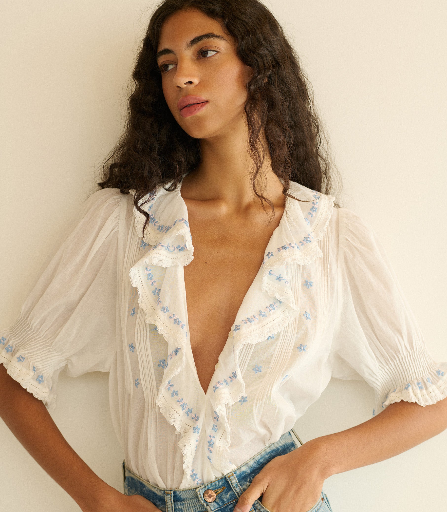 Woman wearing a white blouse with blue floral and blue jeans against a plain background