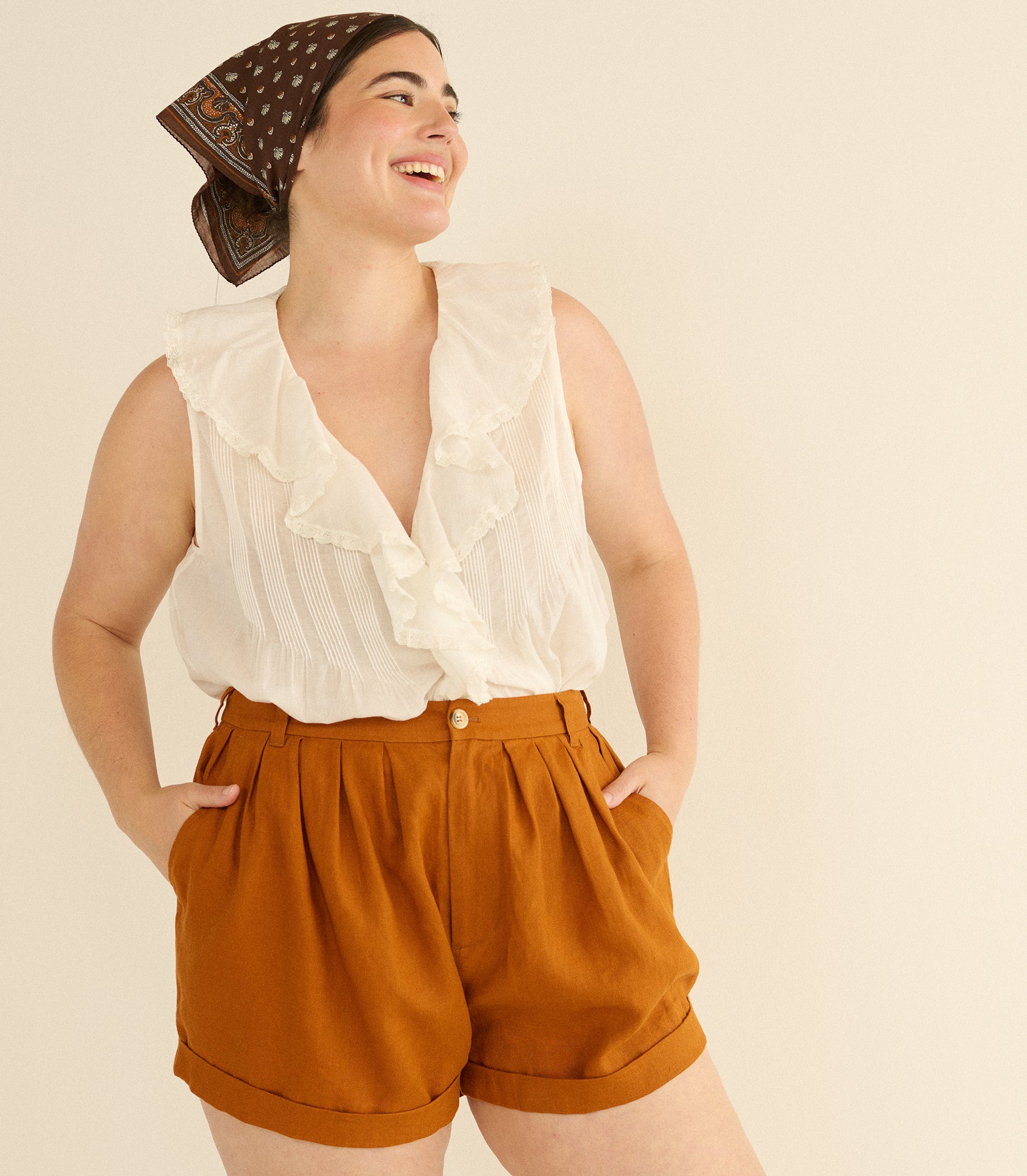 Woman wearing a white blouse and rust-colored shorts with a patterned headscarf on a beige background 4
