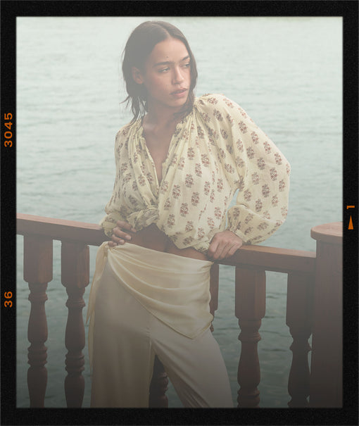 Woman in a floral blouse and white pants standing by a misty water railing.