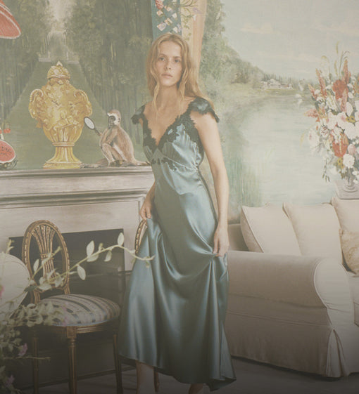 A woman wearing a green satin slip gown with lace trim, standing in a vintage-style room with a sofa, fireplace, and floral wallpaper.